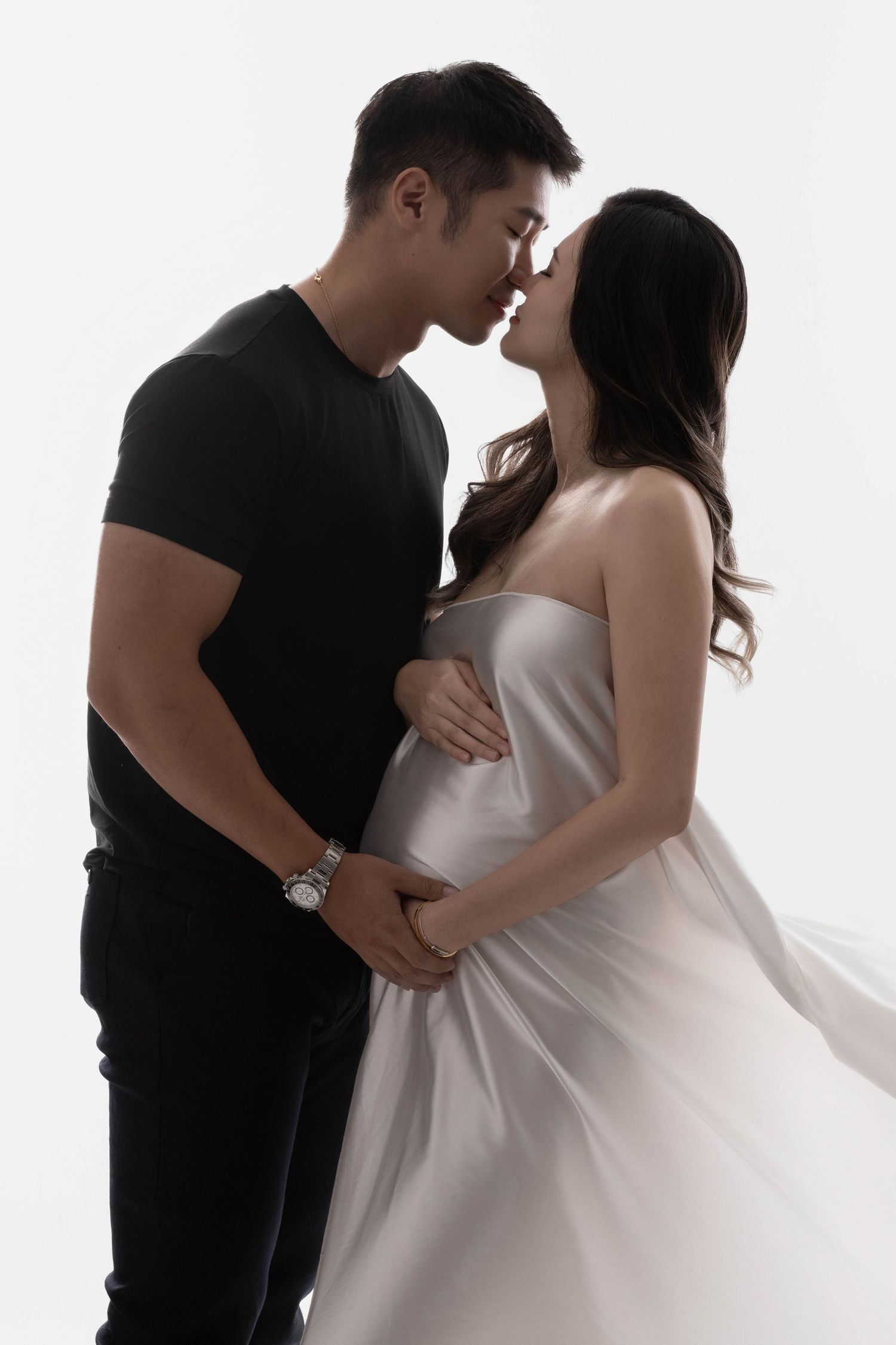Intimate couple's maternity photo session with white dress against light background.