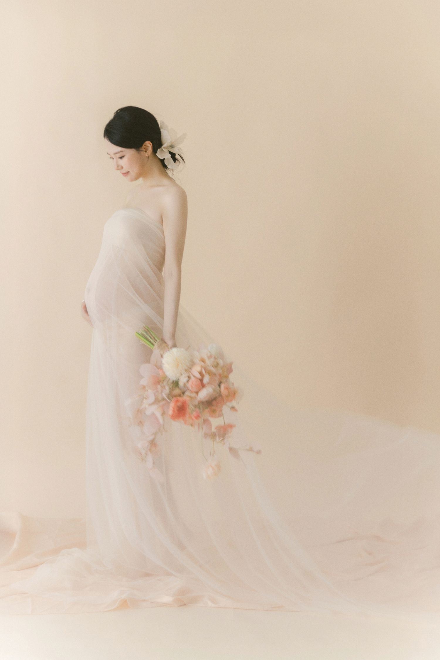 Ethereal maternity photo with soft peach flowers and flowing white fabric against neutral backdrop.