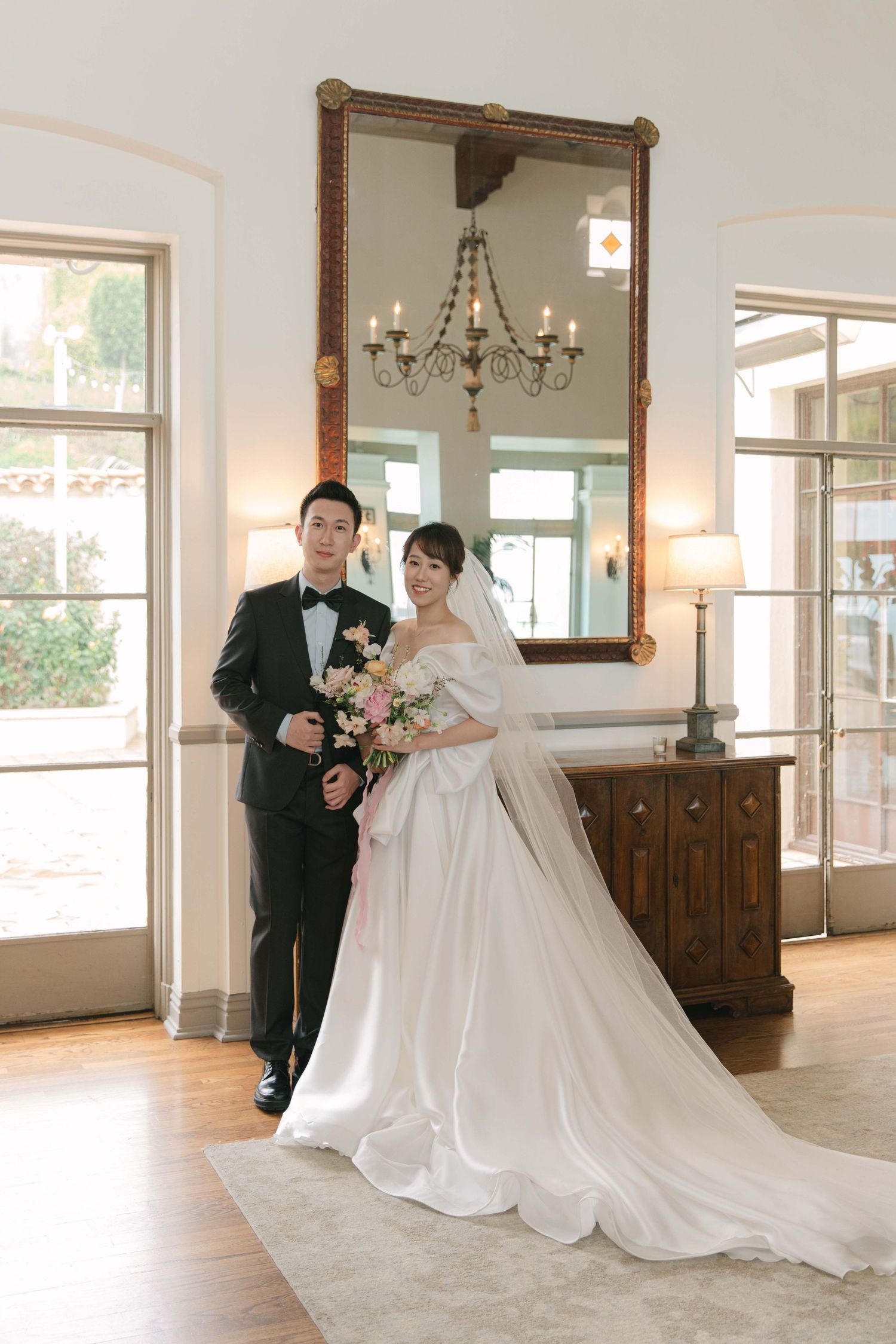 A couple poses in formal wedding attire in a bright, elegant room with wooden furniture.