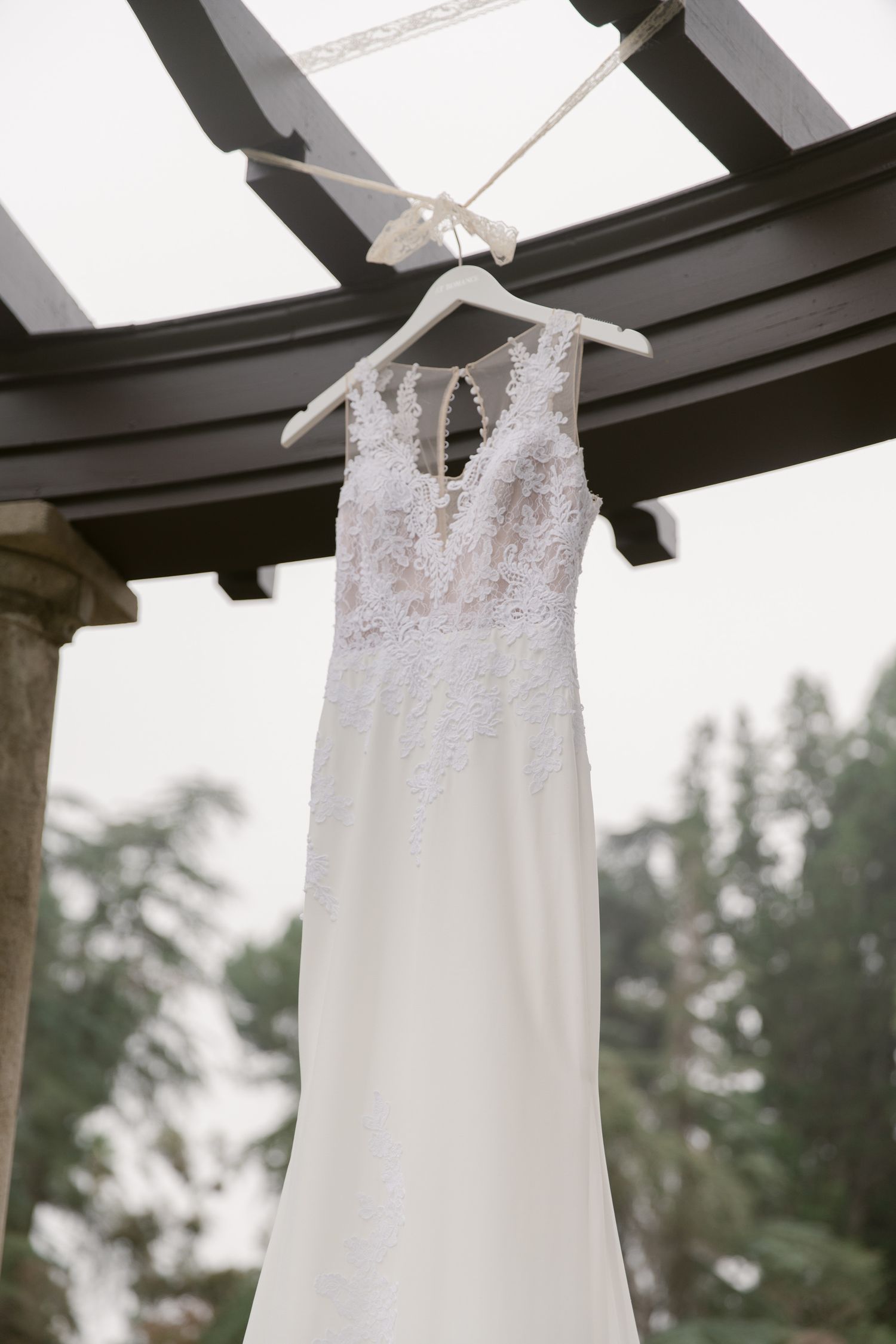 A delicate white lace wedding dress hangs from dark wooden beams against a bright sky.