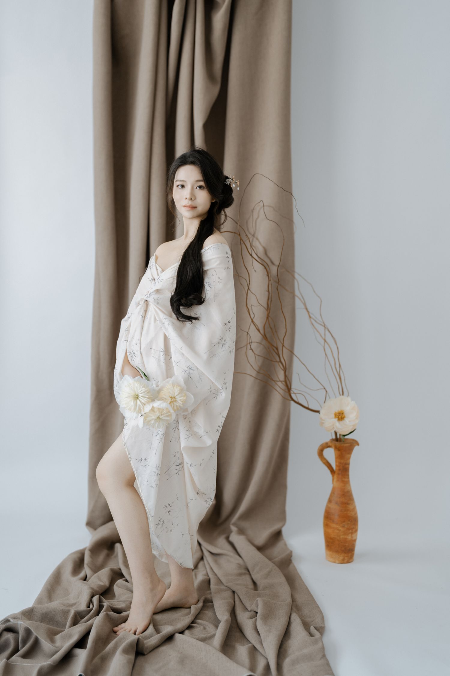 Artistic maternity portrait in flowing white dress against draped backdrop.