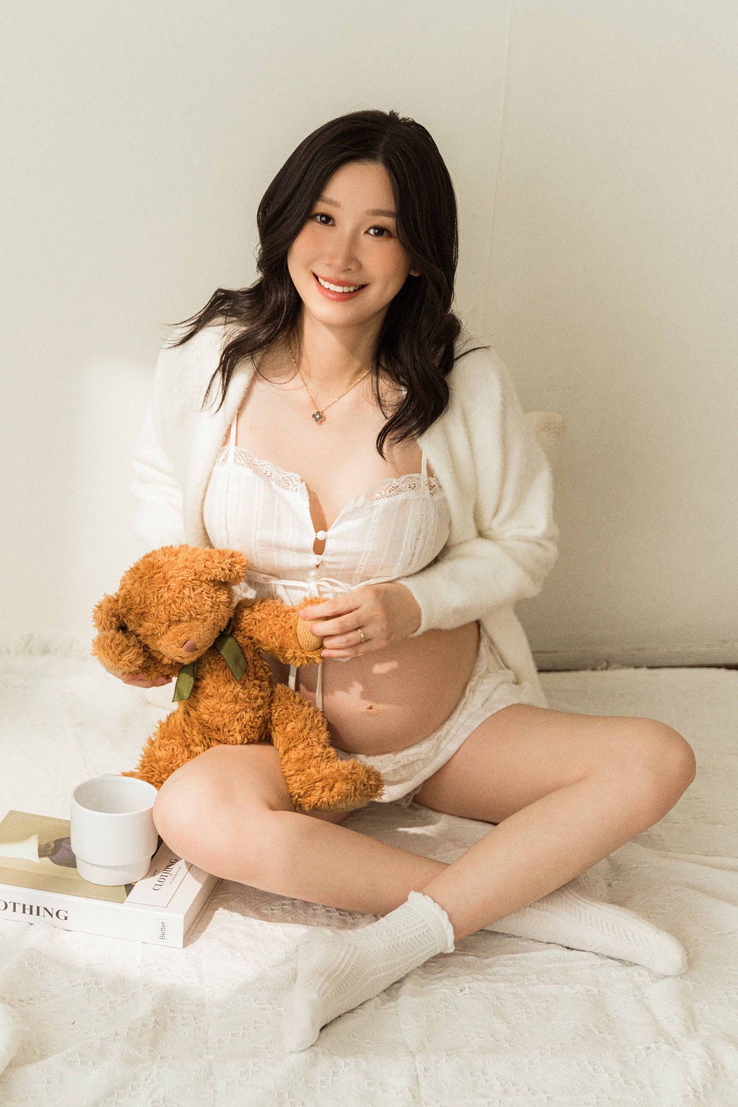 Cozy maternity photos with teddy bear and white cardigan on light bedding.