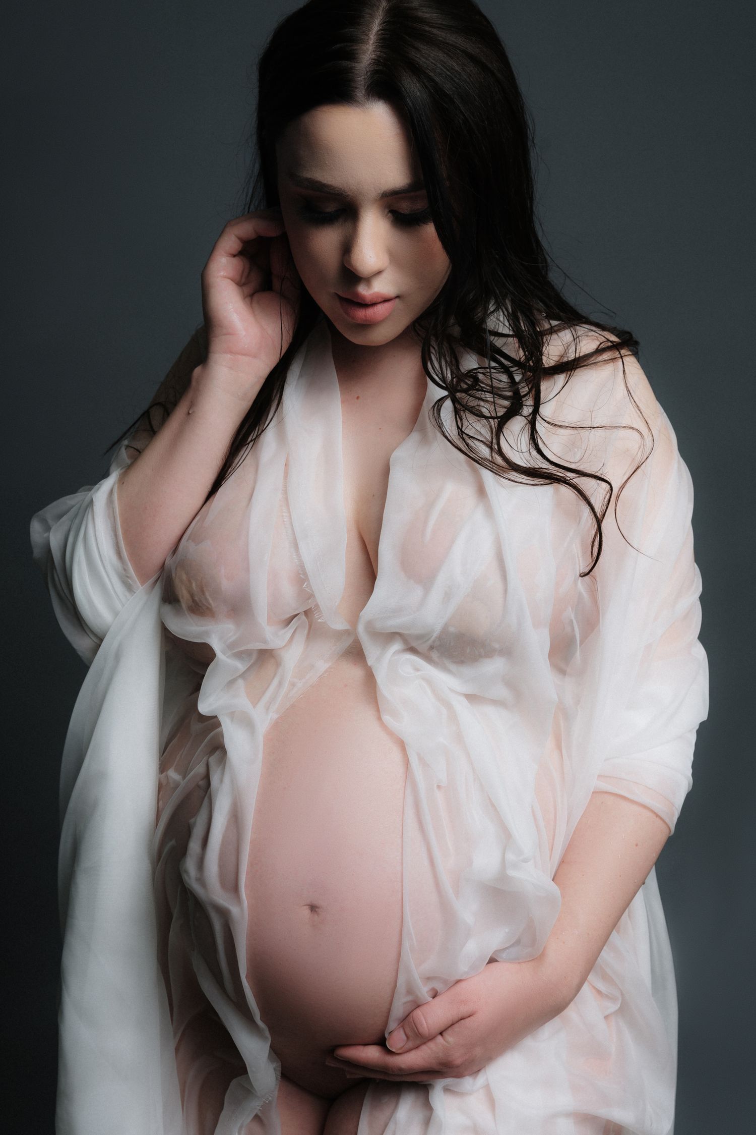 Ethereal maternity portrait in sheer white robe against dark background.