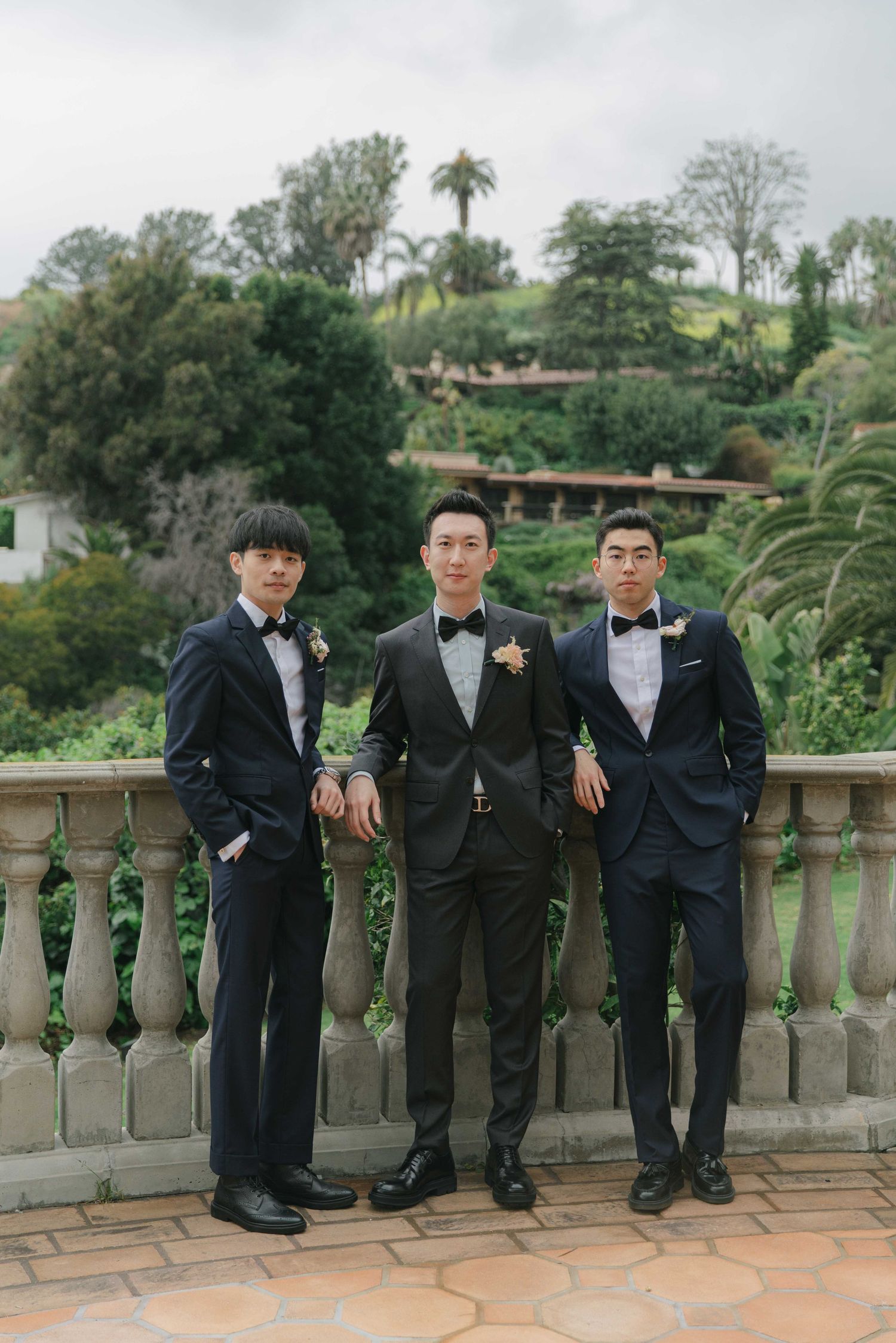 Groomsmen in black tuxedos pose casually against a stone balustrade with palm trees in background.
