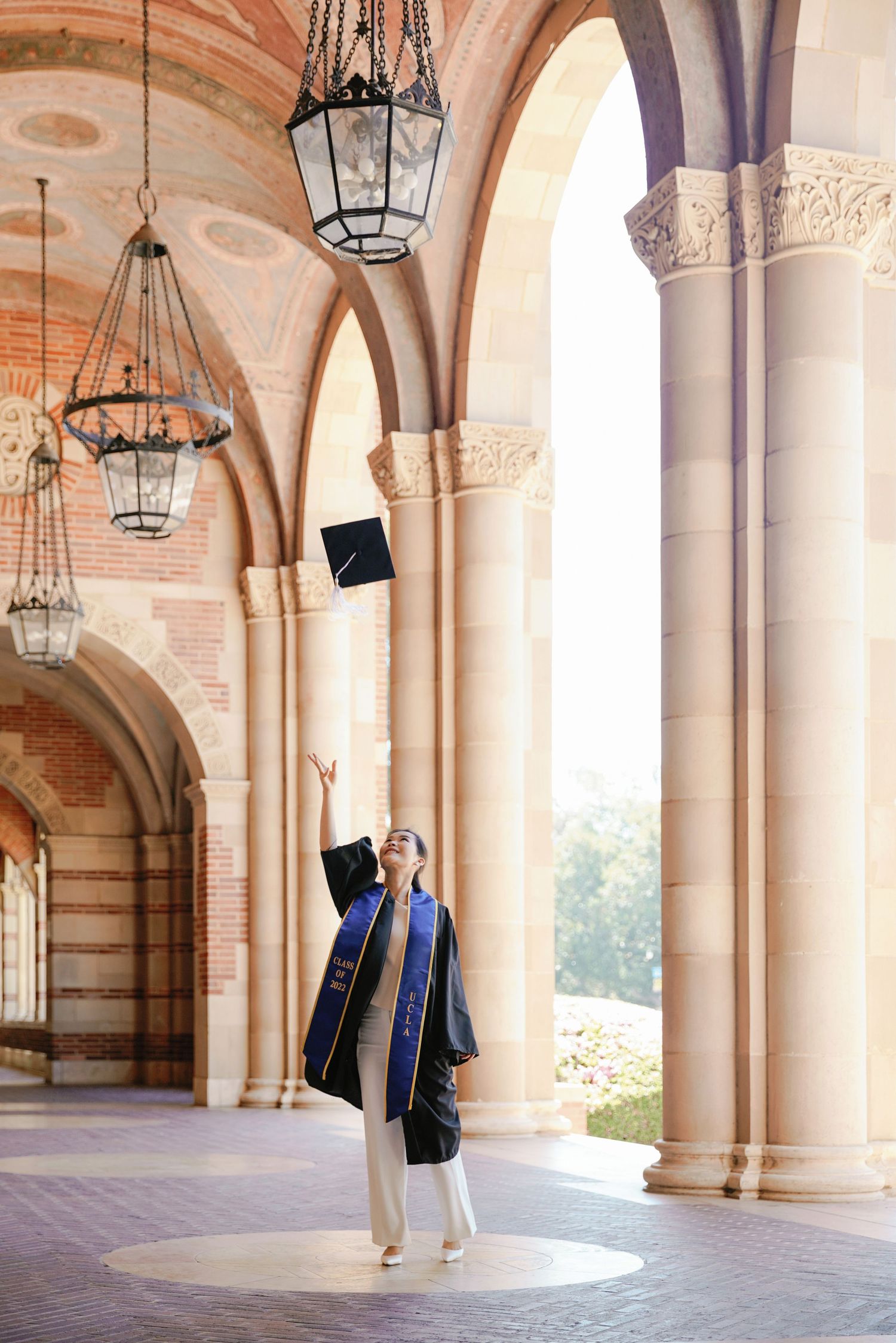Graduates celebrate by tossing caps in ornate arched hallway with hanging lanterns.