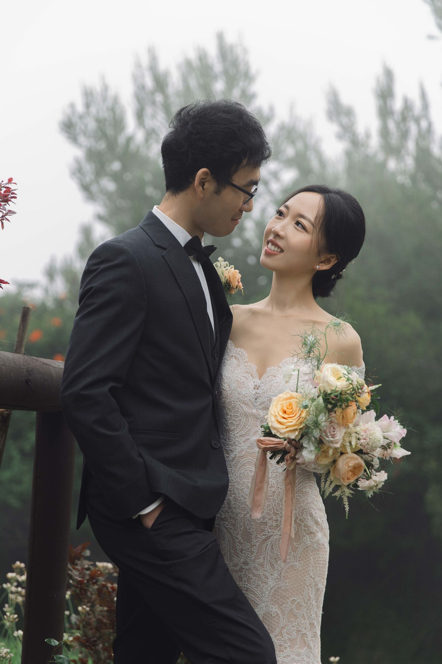Happy couple shares intimate moment in garden while holding peach and cream floral bouquet.