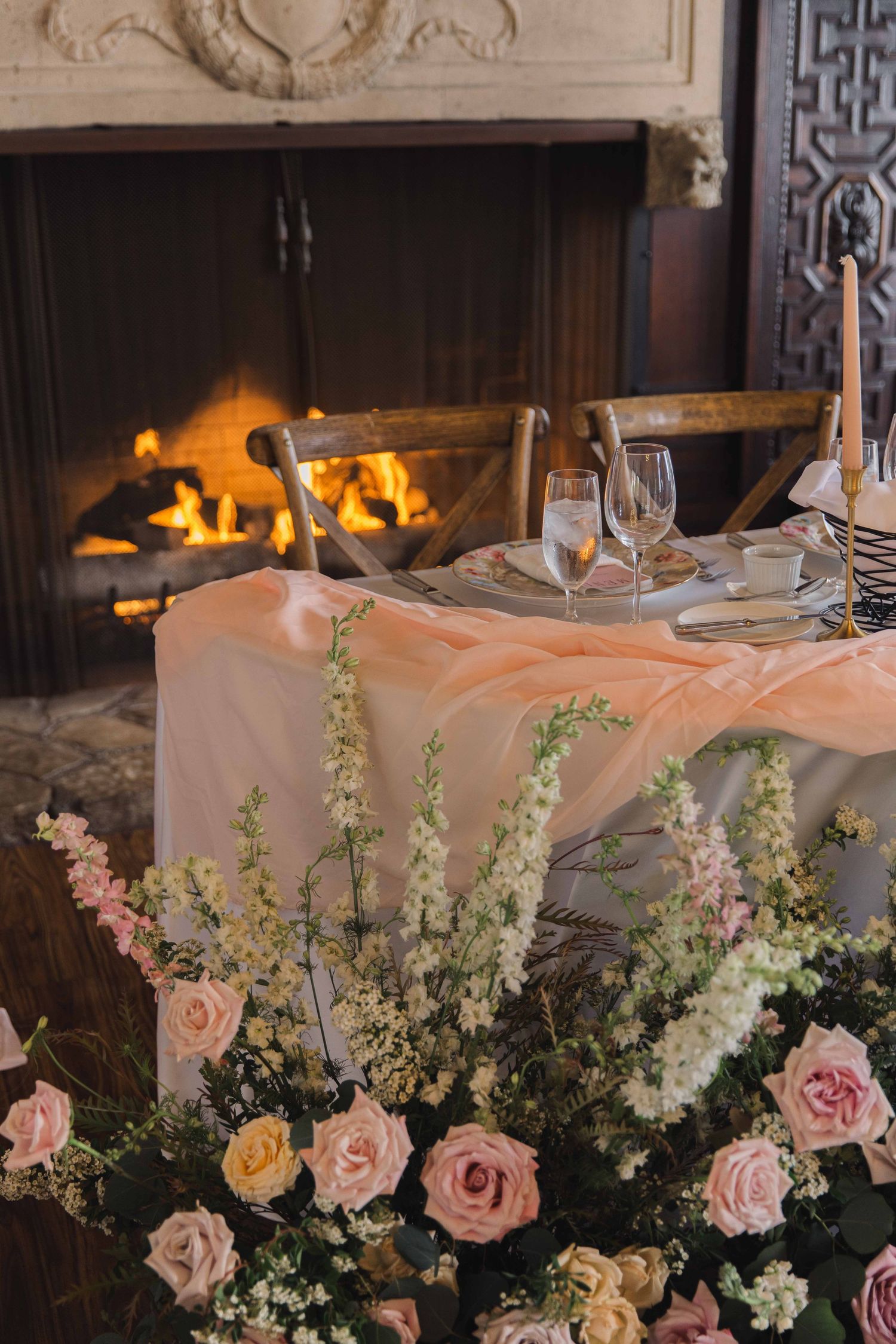 Elegant table setting with pink roses and delicate flowers arranged near a cozy fireplace with glowing flames.