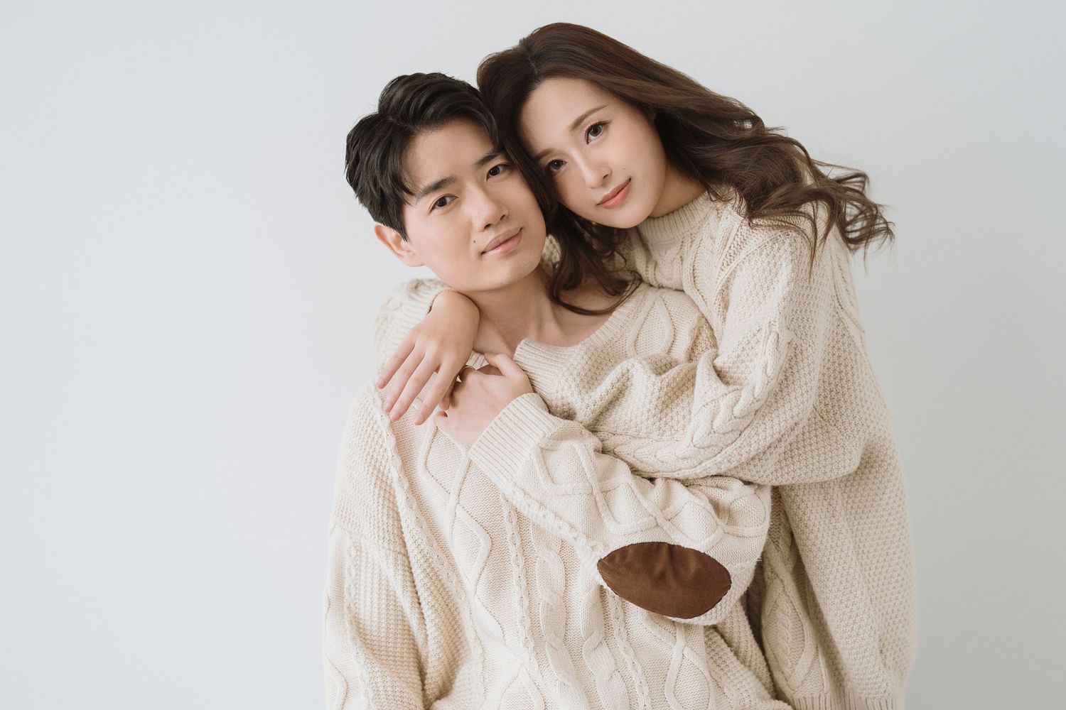 Romantic couple poses together wearing coordinated beige knit sweaters against white background.