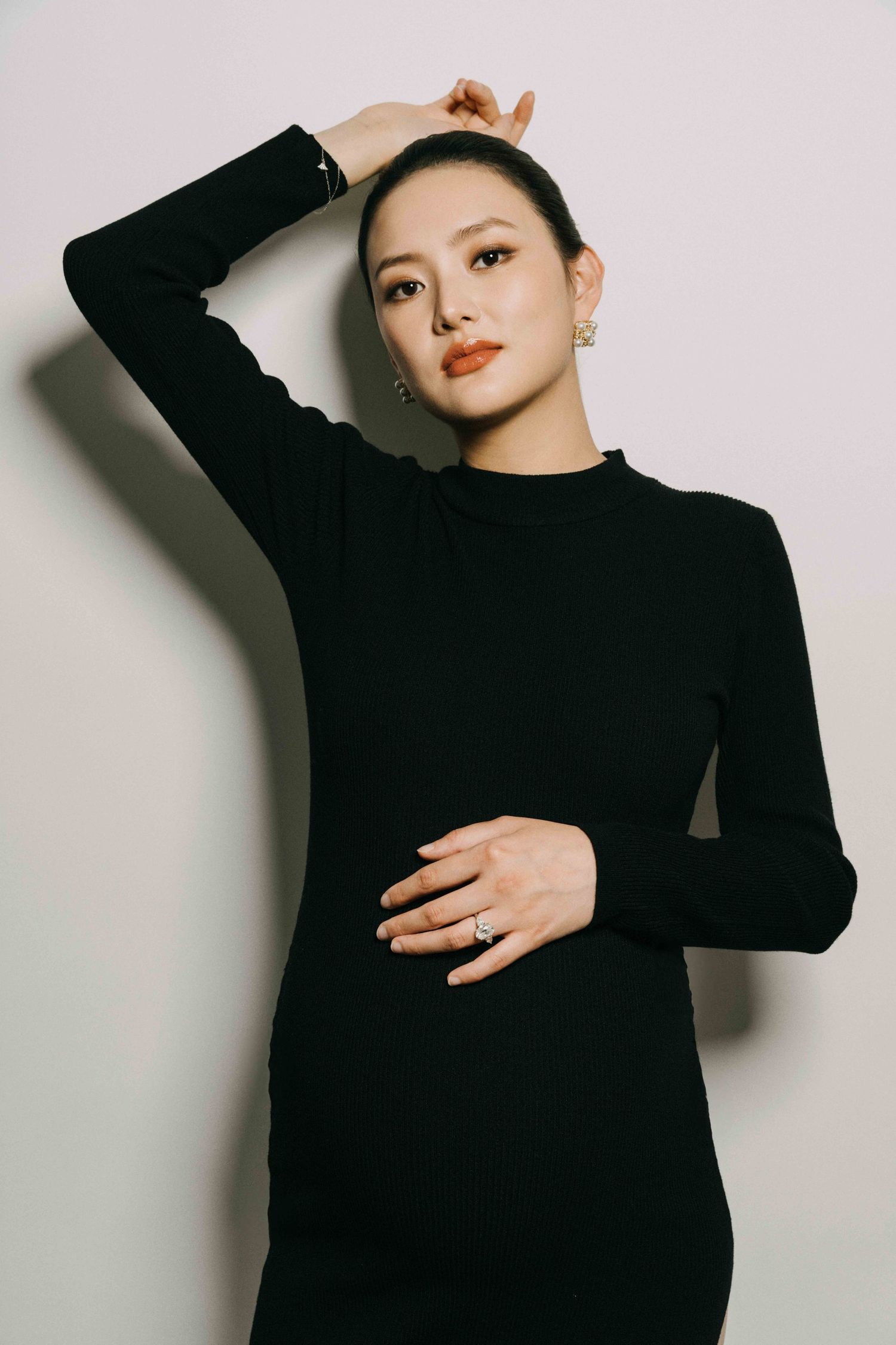 Expectant mother in black turtleneck dress poses elegantly against white wall.