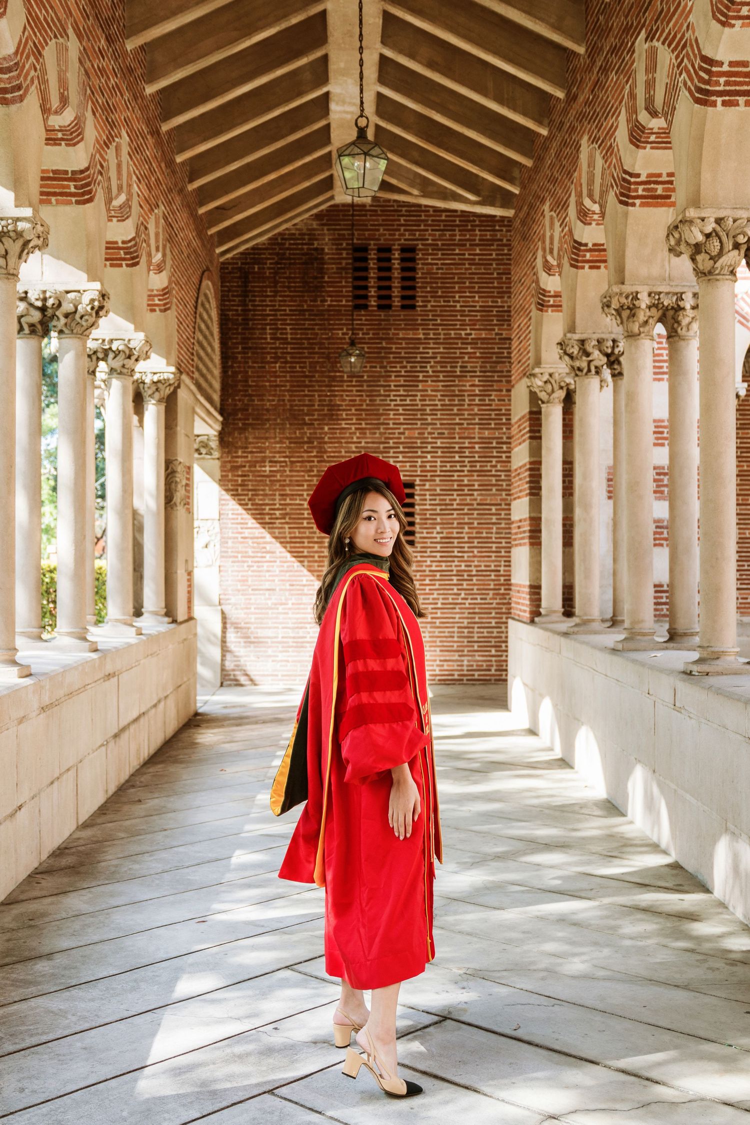 Graduate in red regalia poses in sunlit brick colonnade with white columns.
