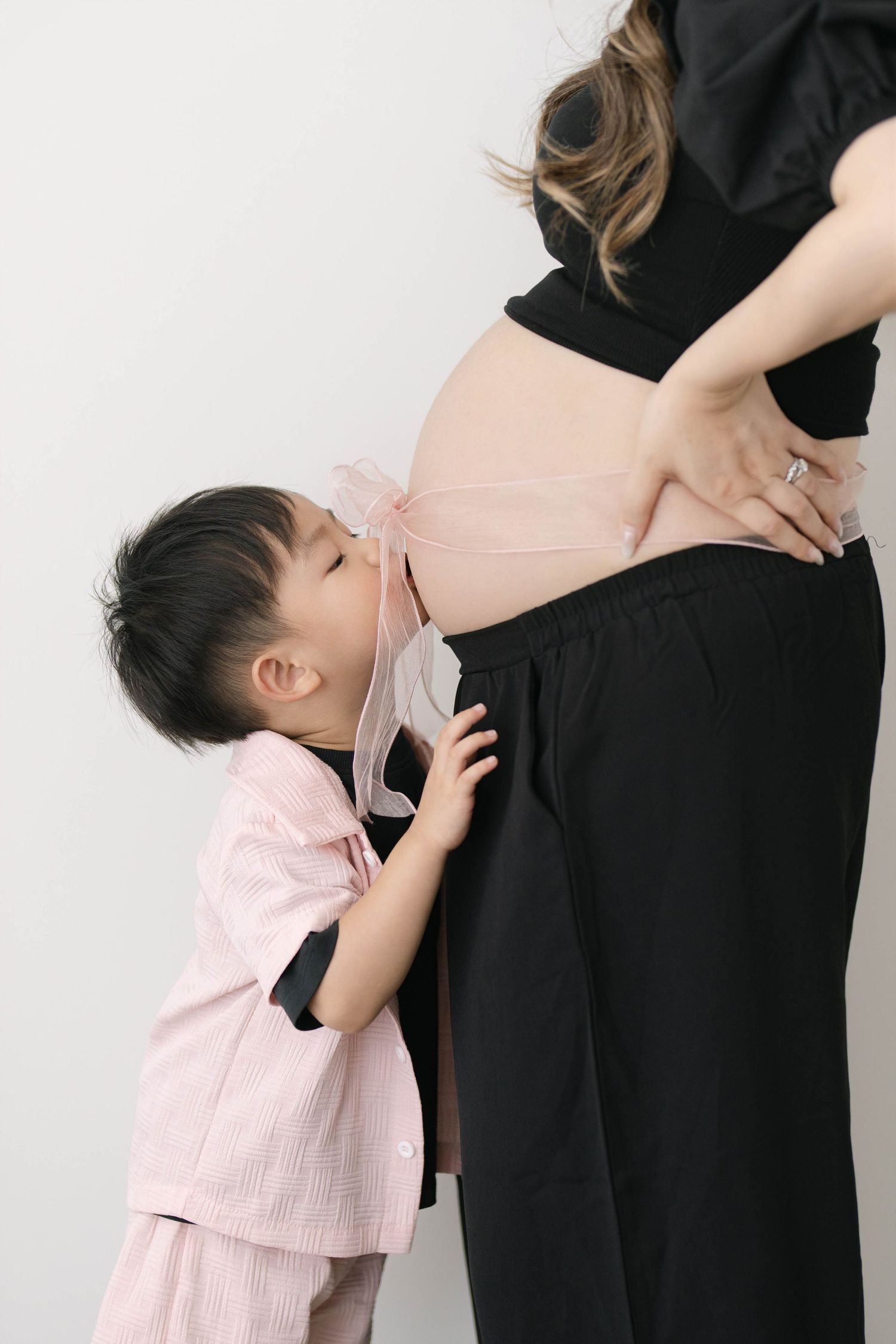 Young child in pink outfit kisses pregnant belly while standing beside mother in black dress.