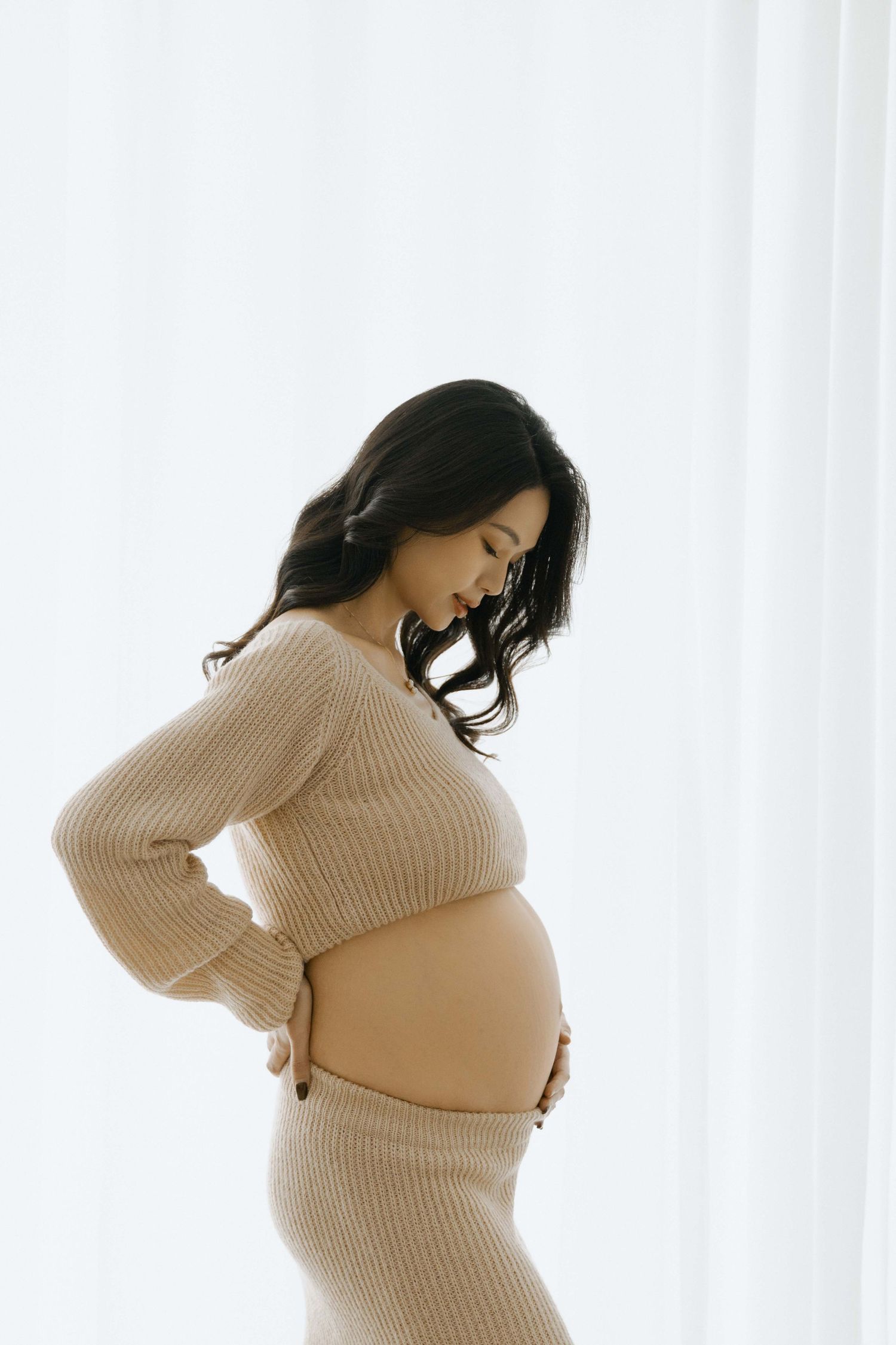 Artistic maternity photo of an expectant mother in a beige sweater against white curtains.