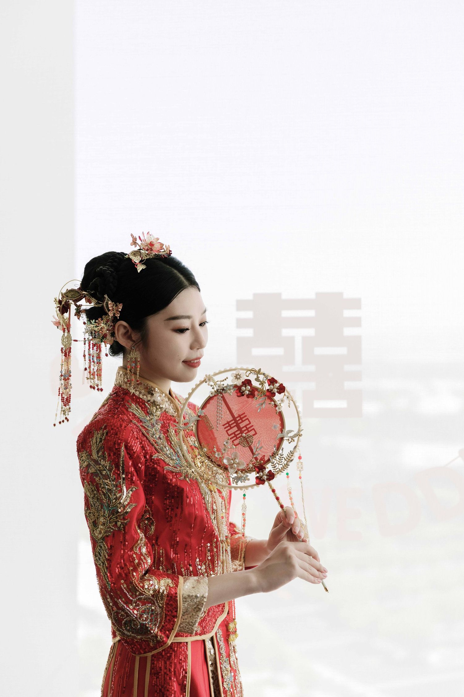 Side profile of person in red Chinese wedding dress holding traditional round fan against white background.