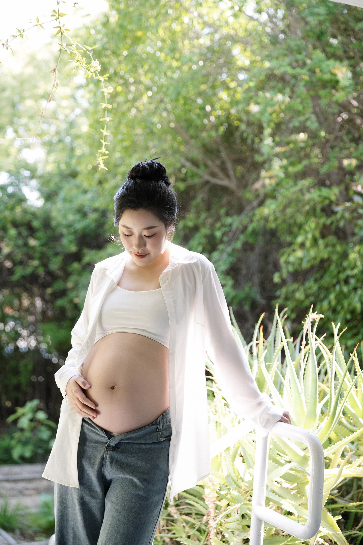 Natural maternity photos in white shirt and jeans amid garden greenery.