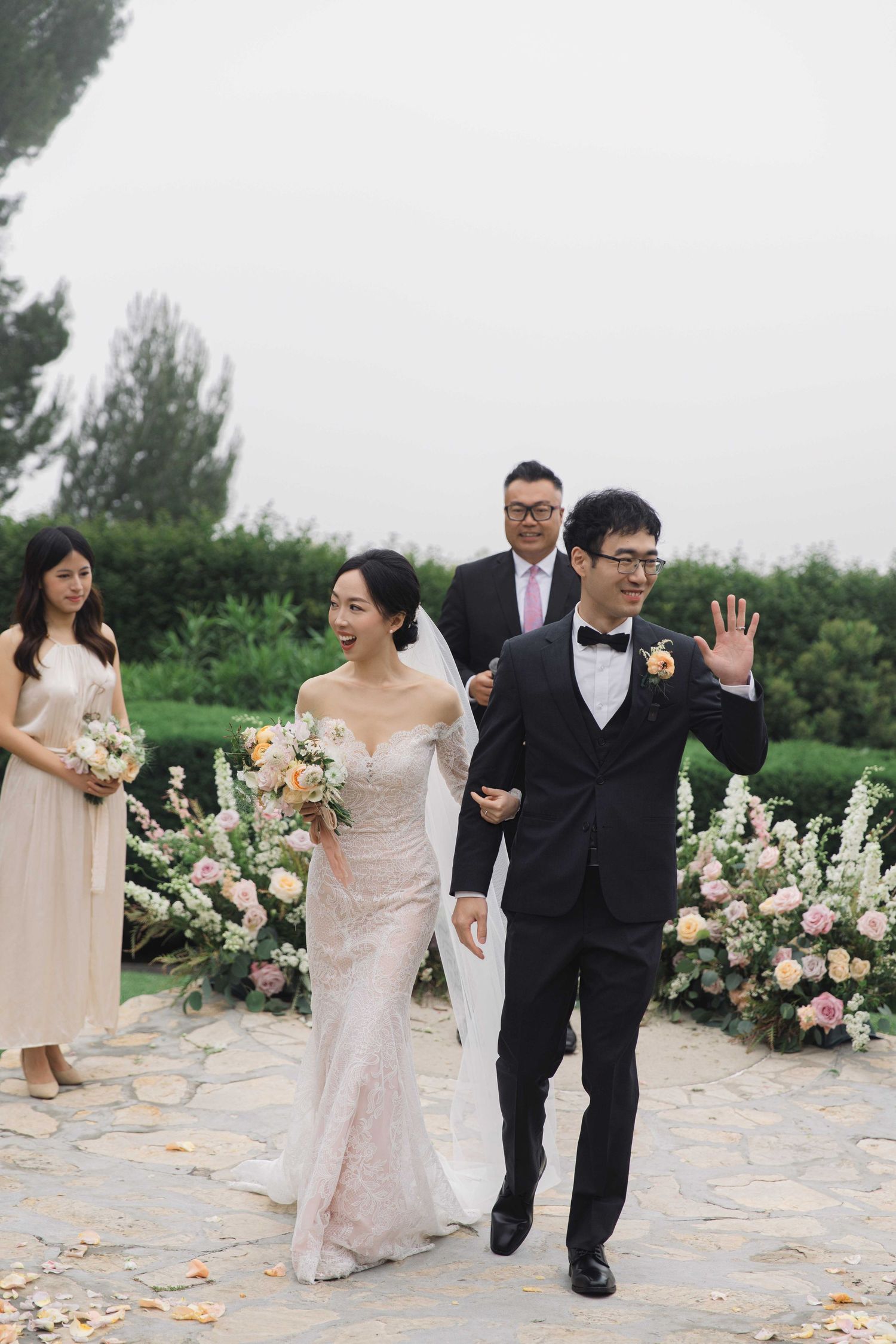Wedding party members walk and celebrate on a stone pathway lined with floral arrangements.