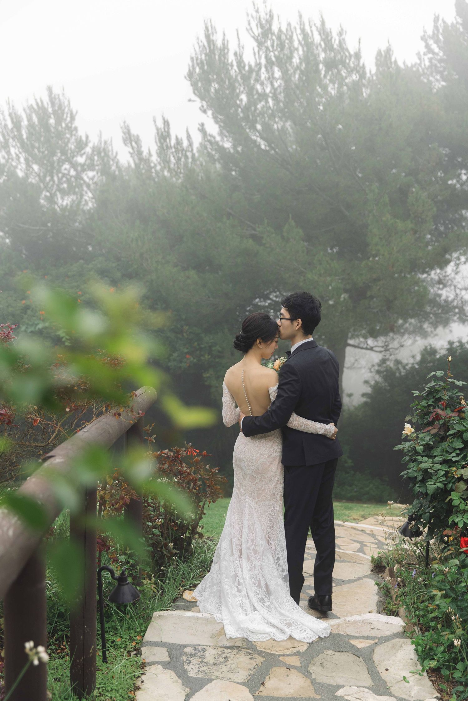 Romantic wedding couple shares intimate moment in misty garden with blooming roses.