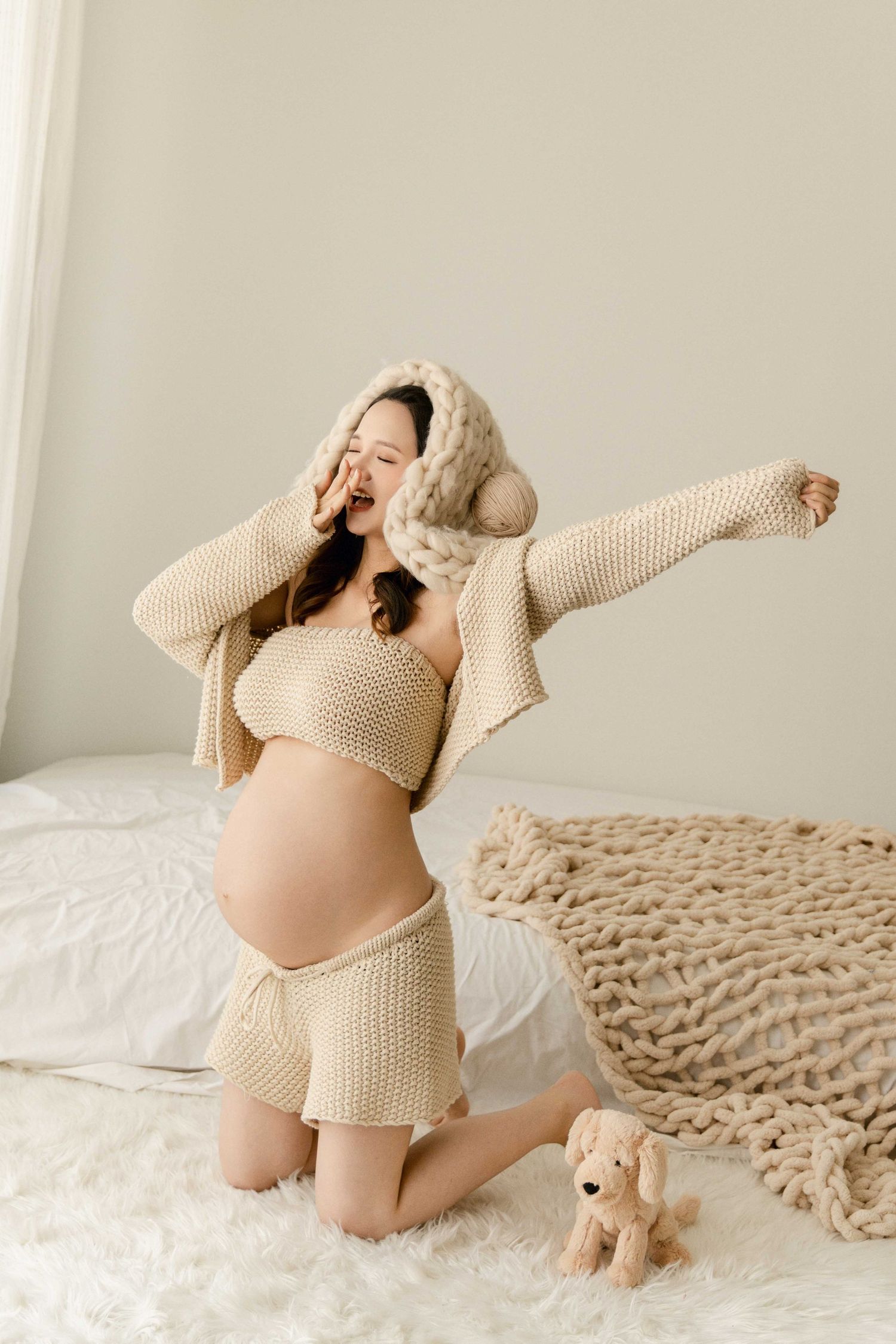 Energetic maternity poses in cream knit outfit with stuffed toy on white bed.