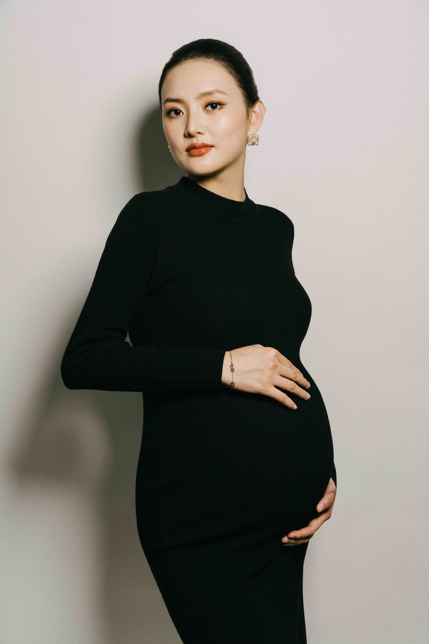 Stylish maternity portrait featuring woman in fitted black dress against neutral background.