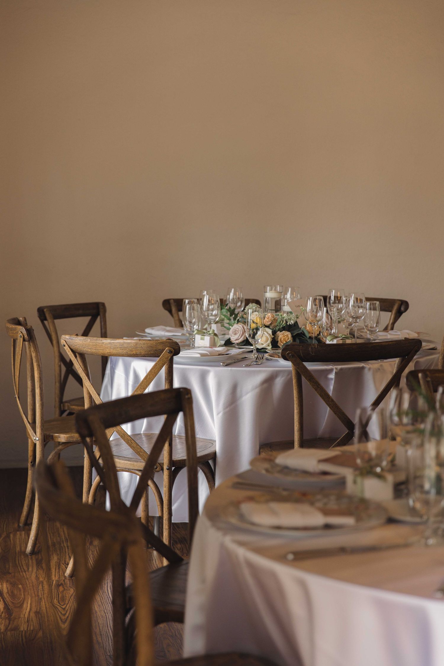 Elegant wedding reception table setup with rustic wooden chairs and white tablecloths in a neutral setting.