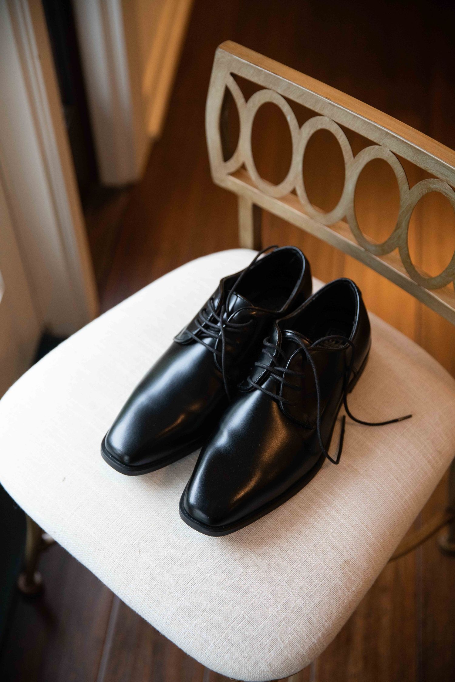 Black leather dress shoes placed on beige upholstered chair with decorative metal backrest.