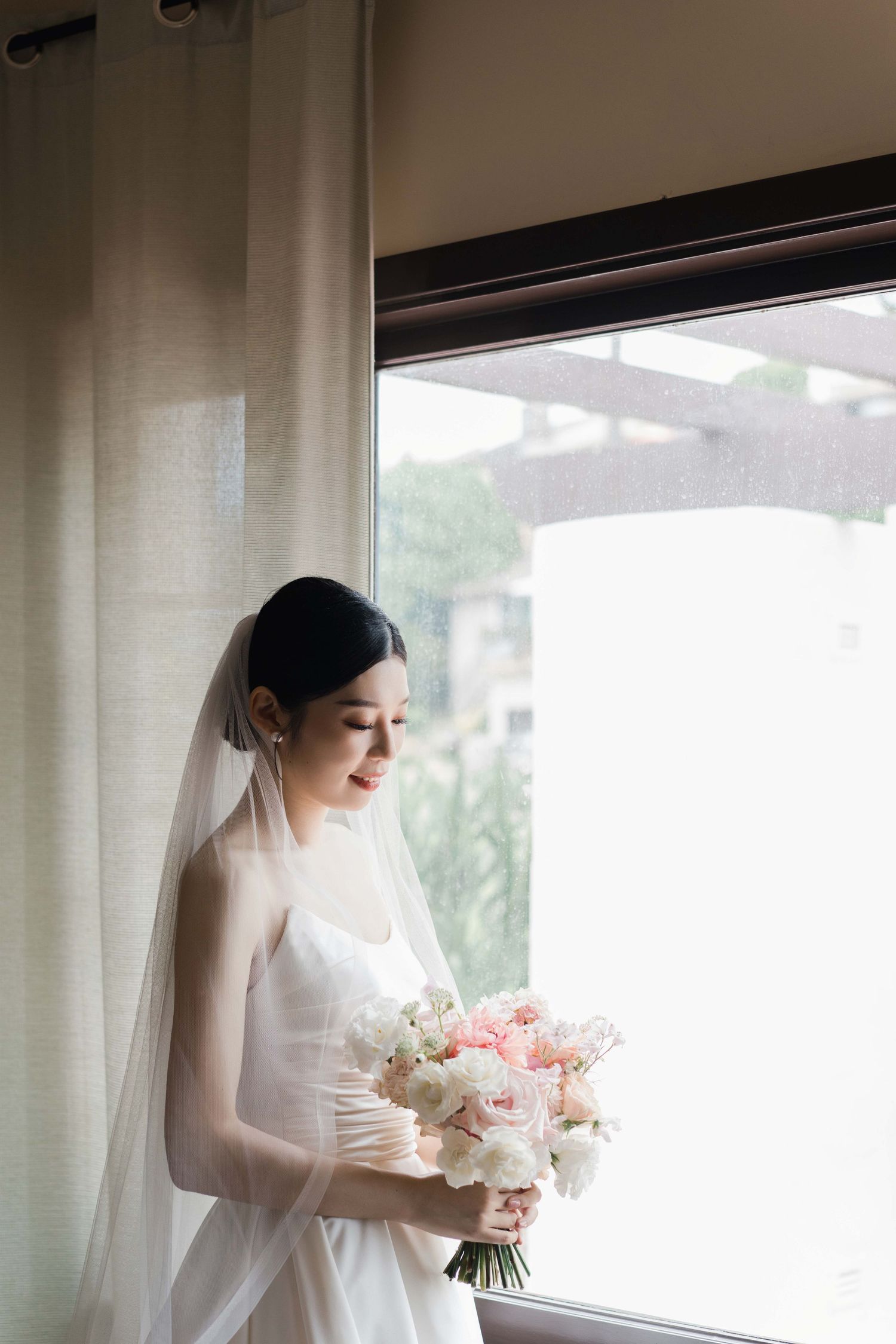 A bride in a white wedding dress holds a delicate pink and white bouquet while standing by a window.