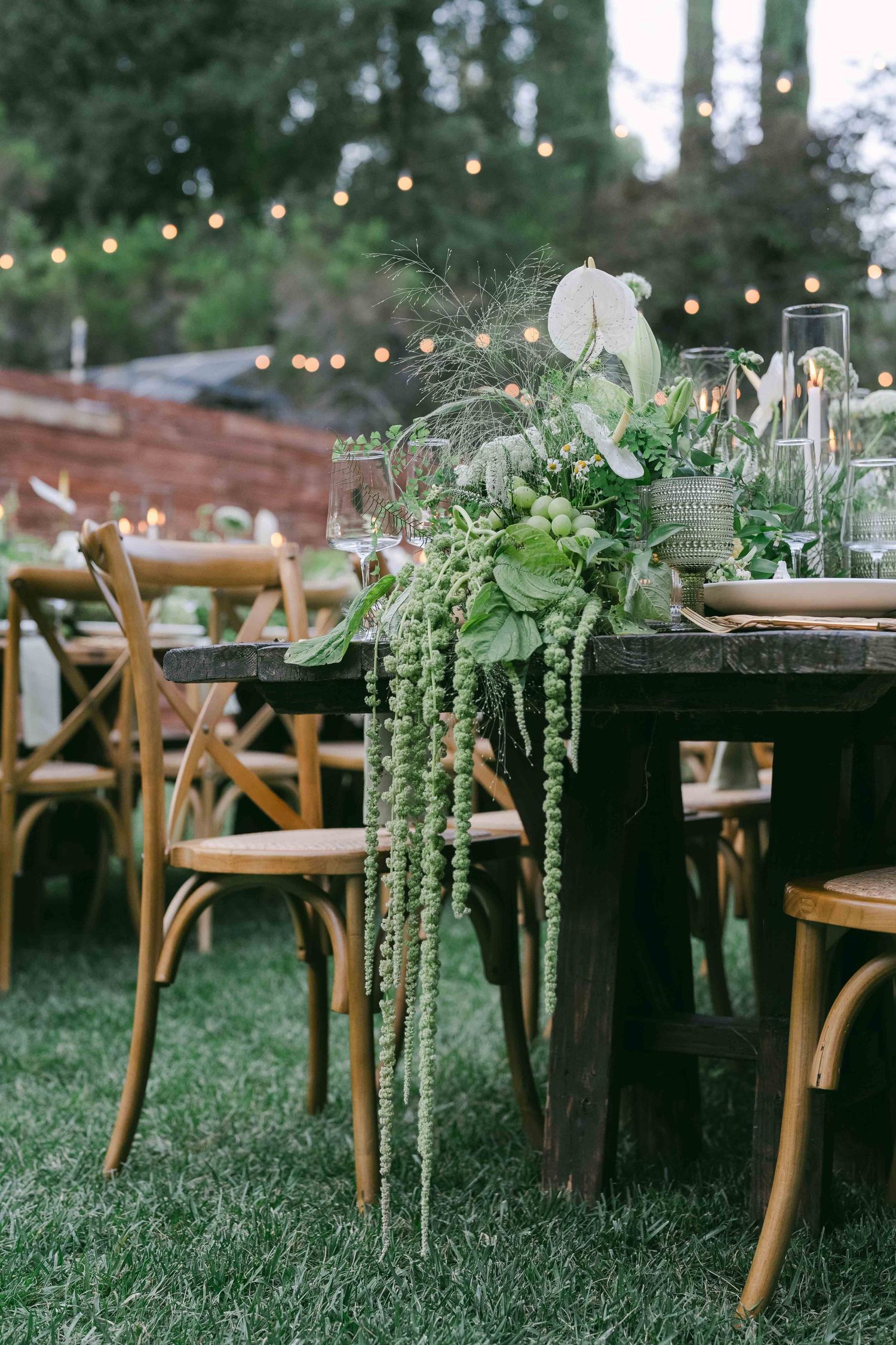 Rustic outdoor wedding reception setup with wooden chairs and hanging string lights.