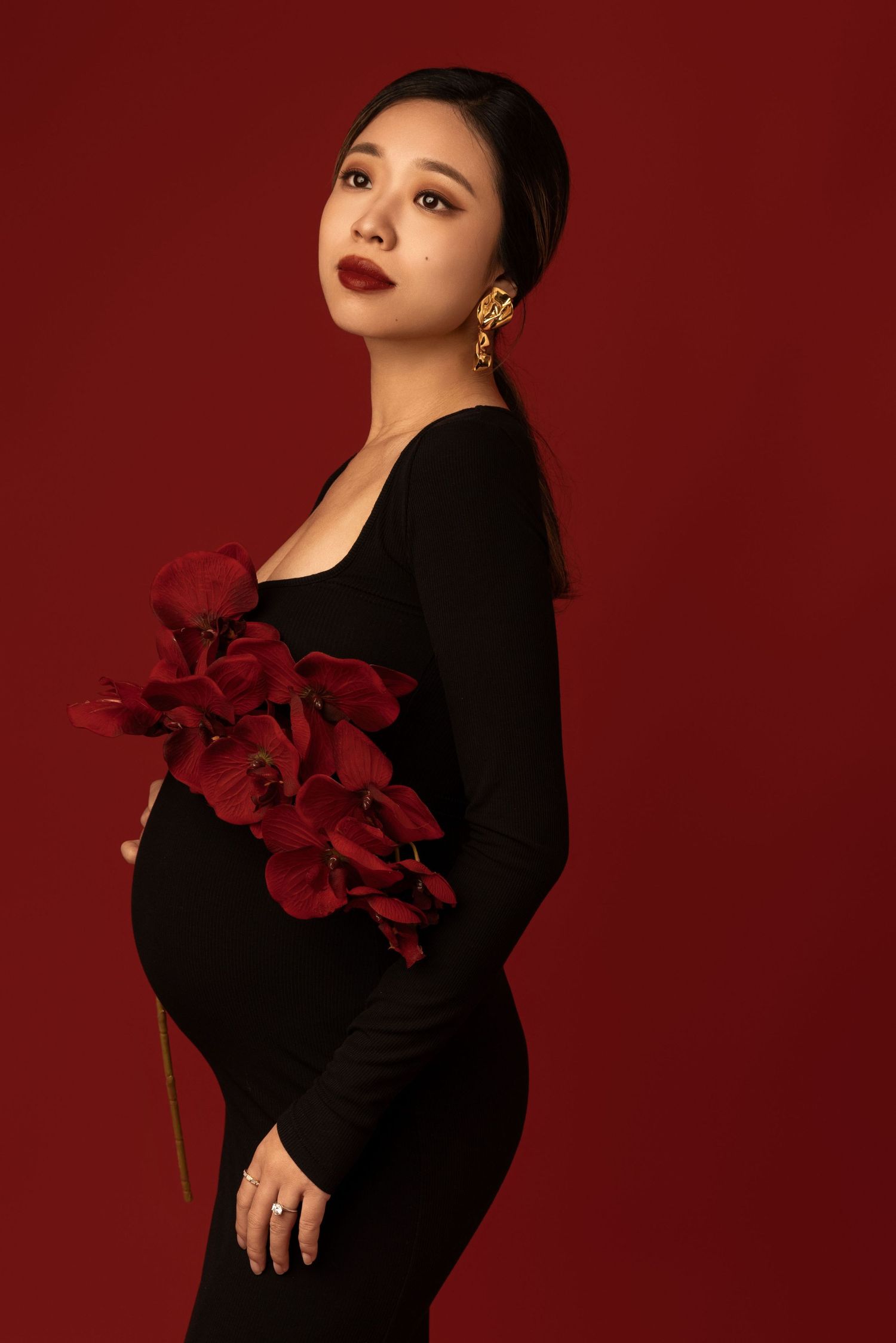 Dramatic portrait in black dress with red orchids against burgundy backdrop.