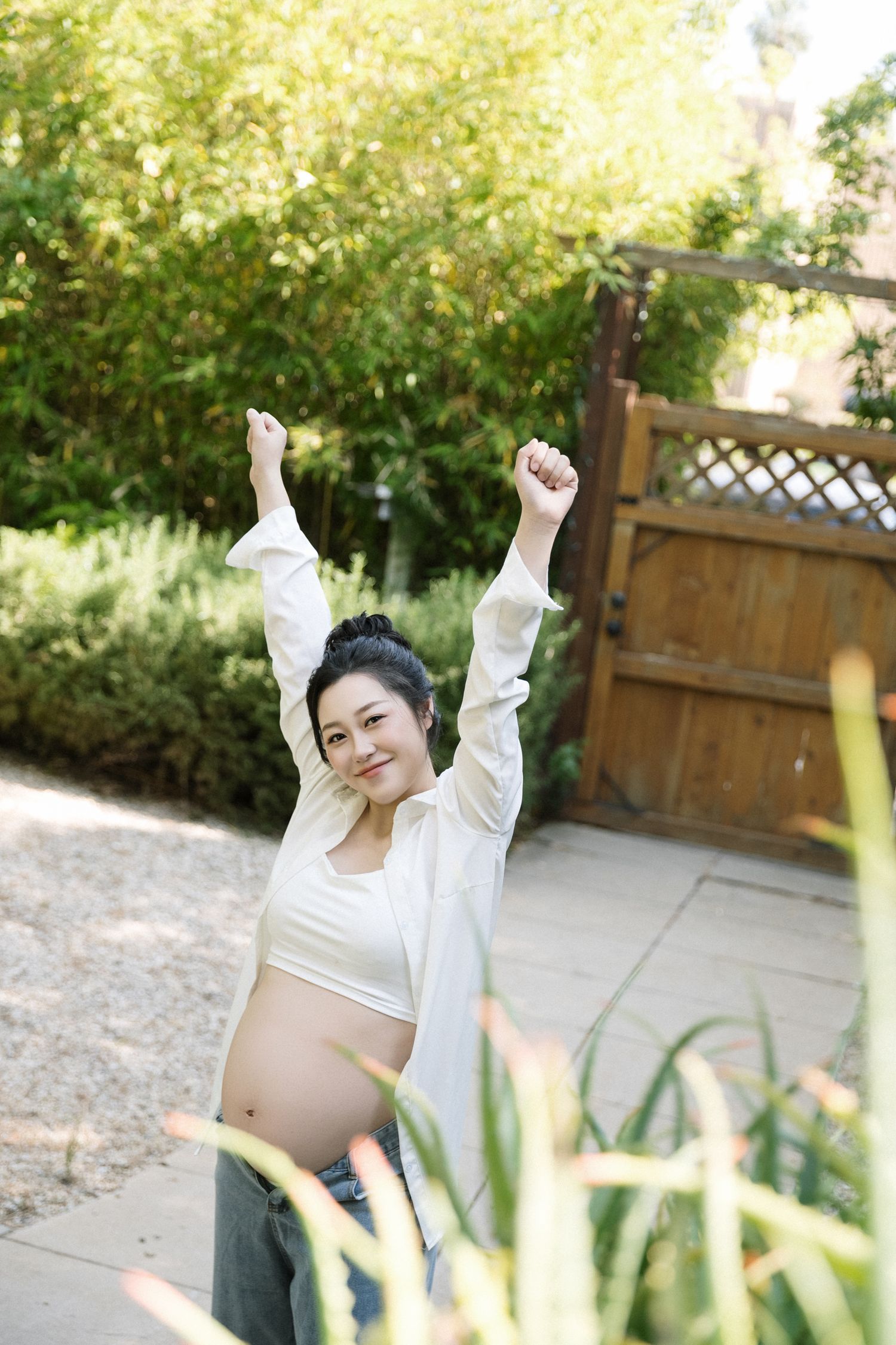 Joyful outdoor maternity photos with raised arms in sunlit garden setting.