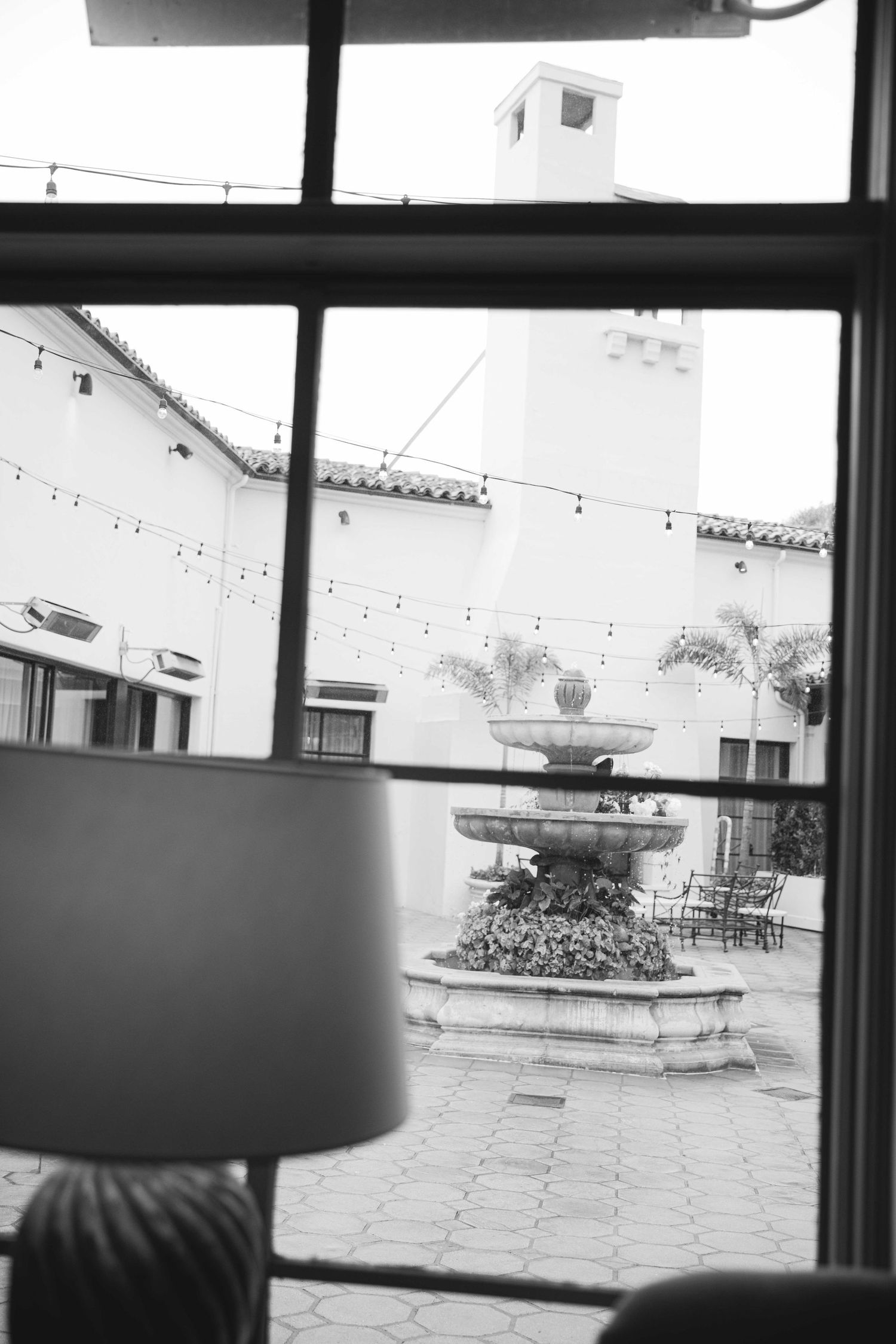 Black and white view through window showing courtyard with string lights and fountain.