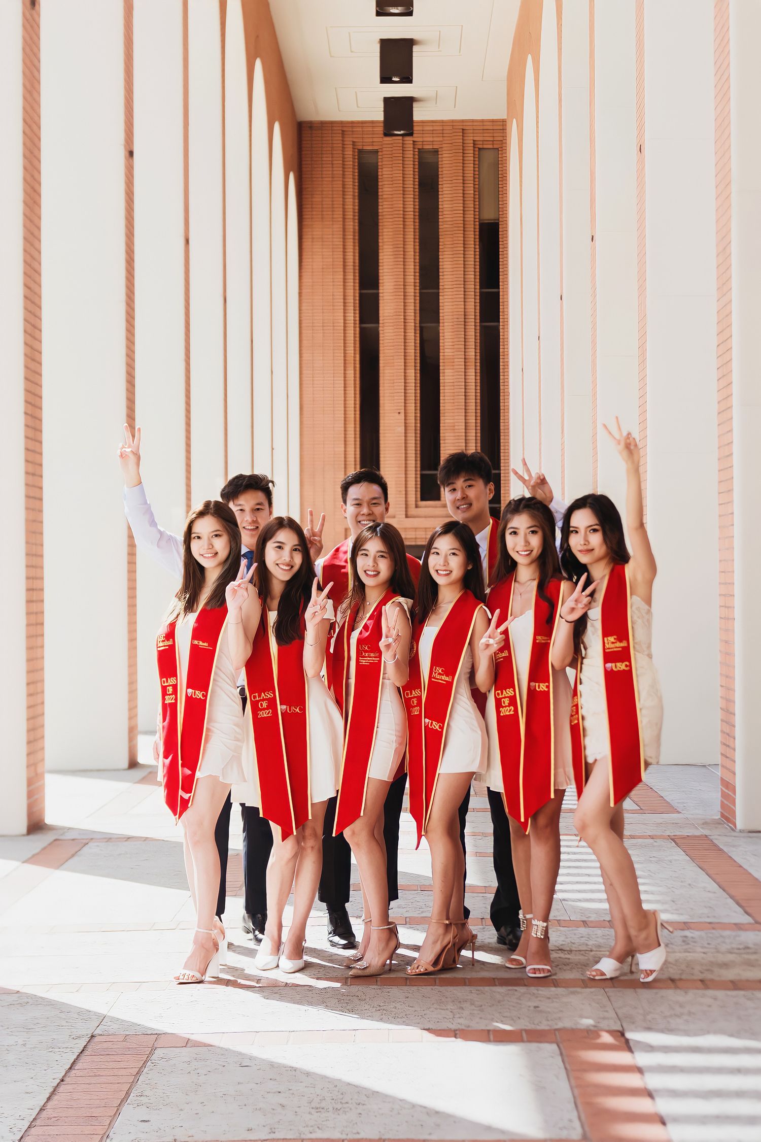 USC graduates in red stoles posing together in a colonnade hallway.