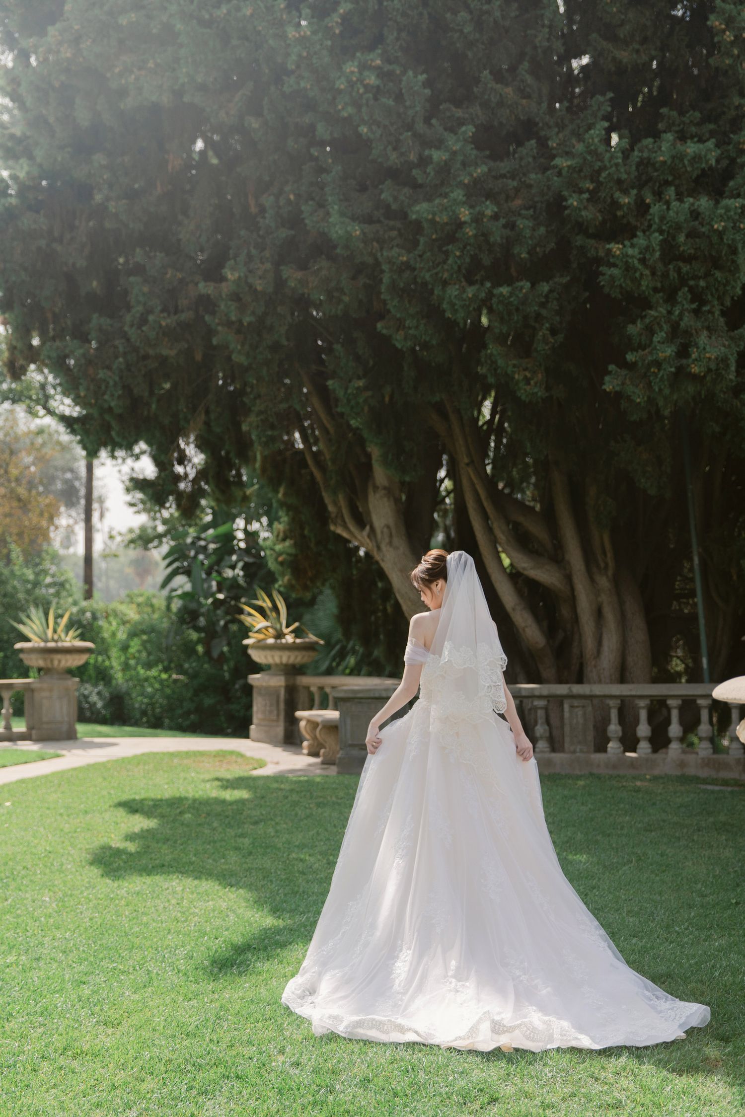Flowing white wedding dress photographed in a serene garden setting with mature trees.