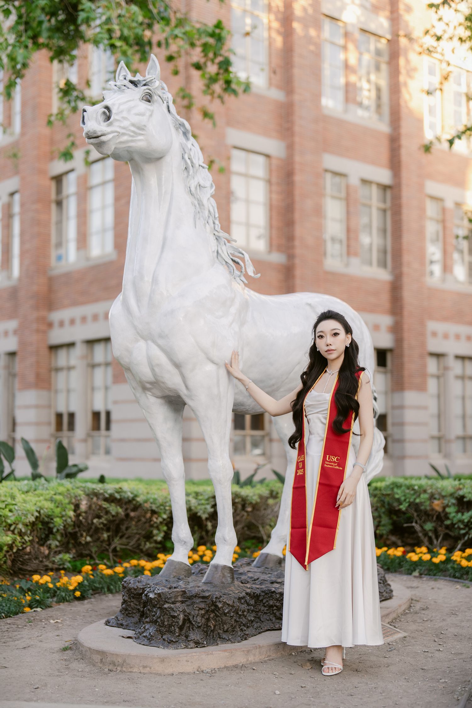 Graduate in white dress and red stole poses with decorative white horse statue on campus.