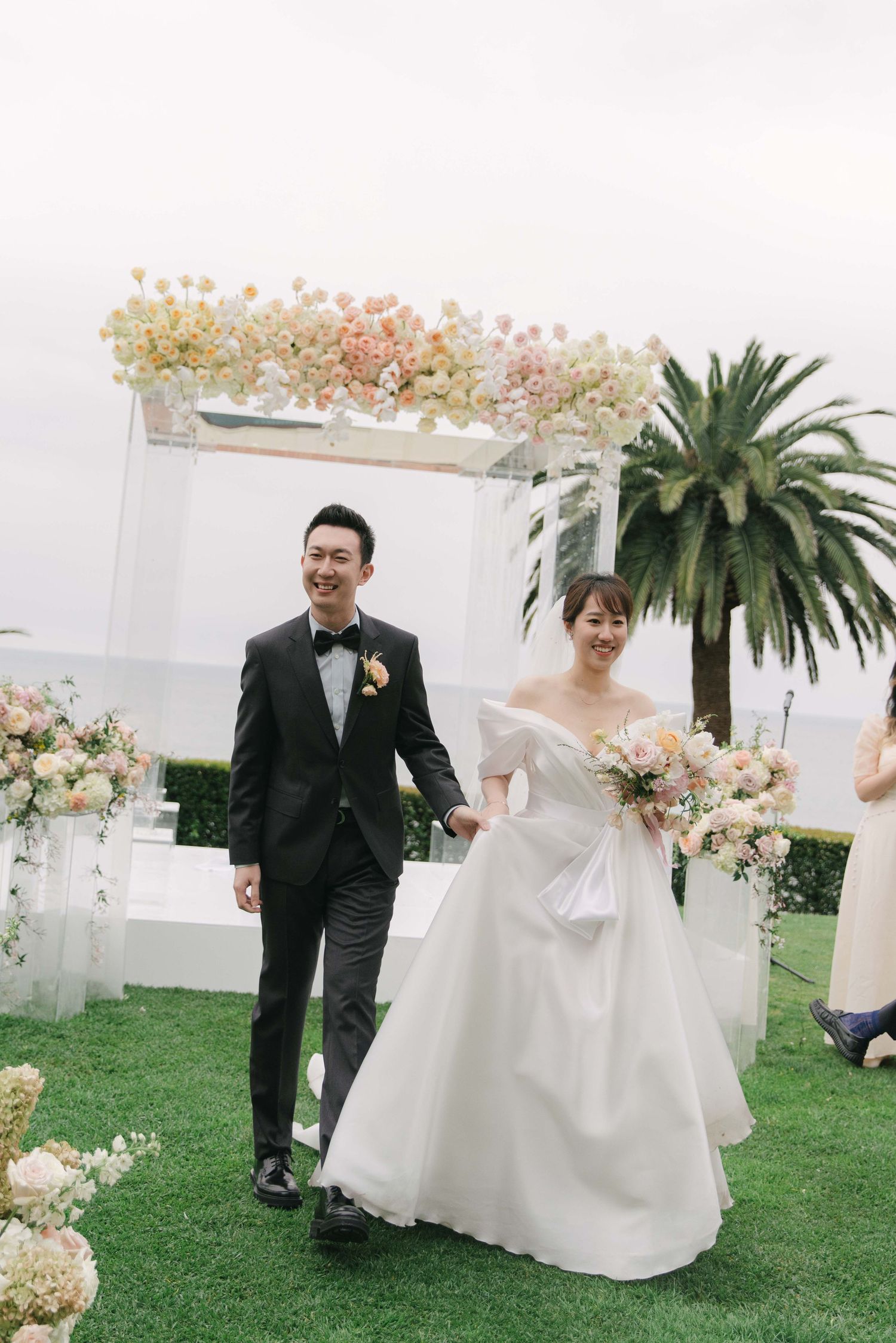 Wedding couple walking on manicured lawn with decorative white arch and palm trees.