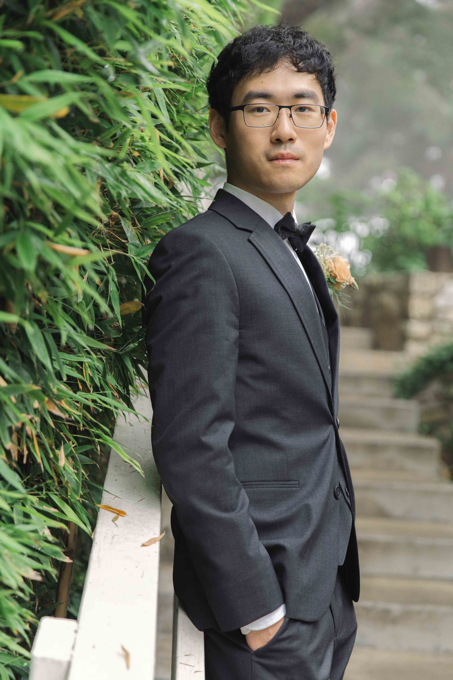 Groomsman in black suit with boutonniere poses against garden foliage.