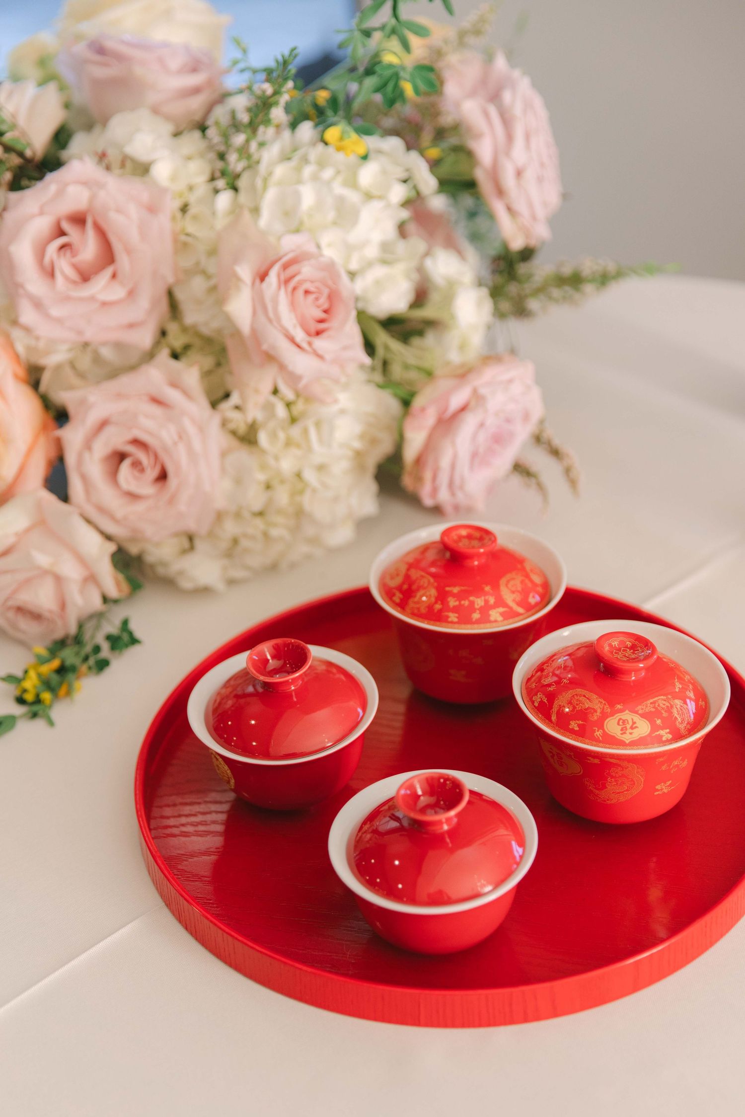 Red ceramic tea cups on a red tray with pink and white floral arrangements in the background.