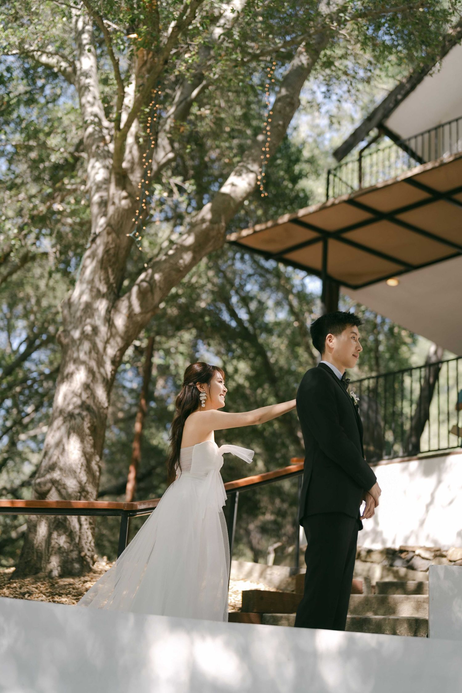 Wedding couple in formal attire standing on a white walkway surrounded by trees.