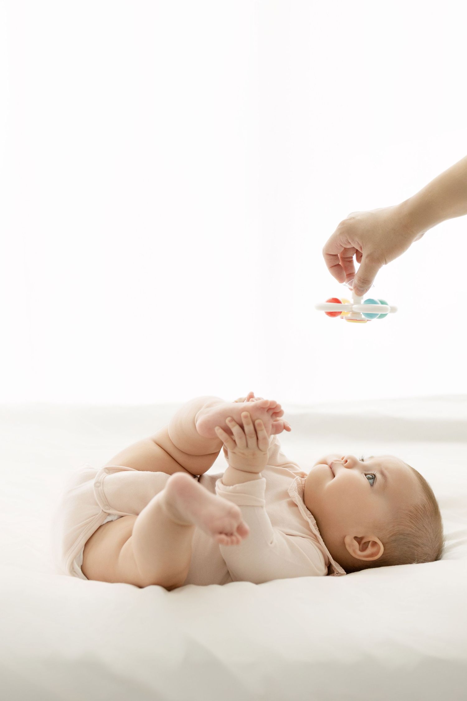 Baby lying on white background reaching for colorful toy during photoshoot.