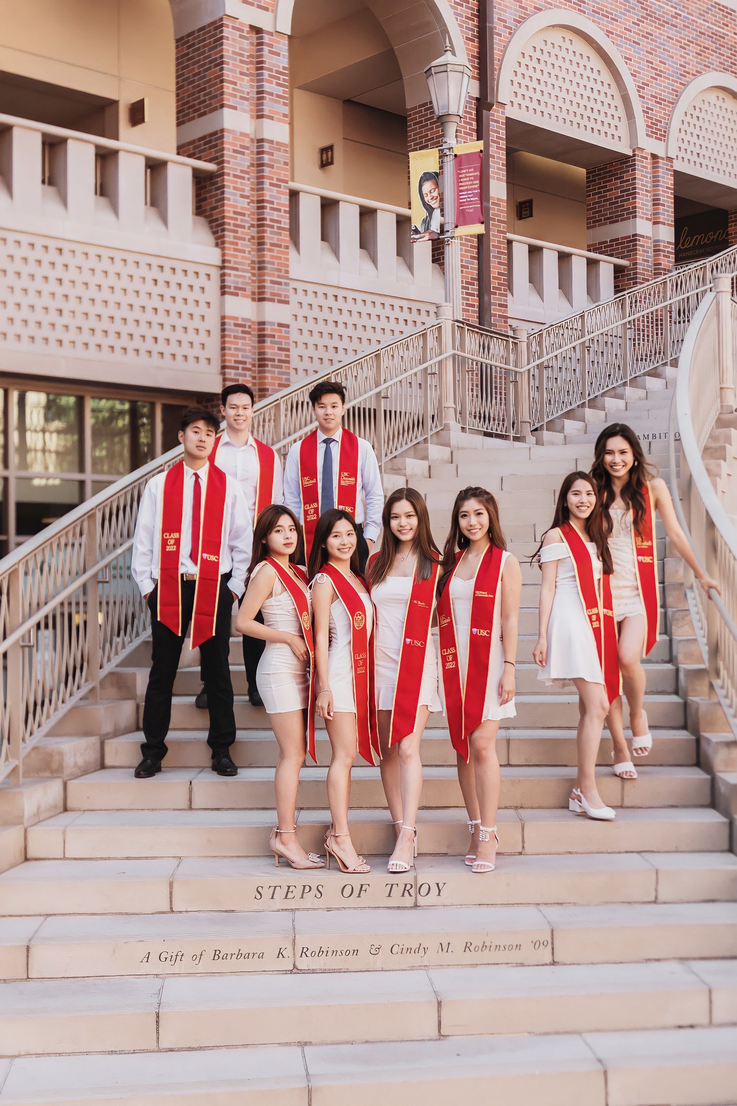 Group of graduates in red USC stoles posing on brick stairs for graduation photos.
