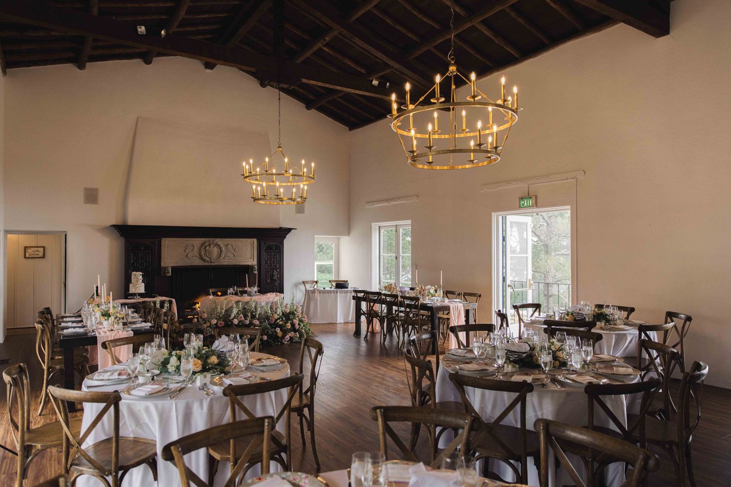 Rustic wedding reception venue with wooden cross-back chairs and elegant chandeliers in a bright open space.