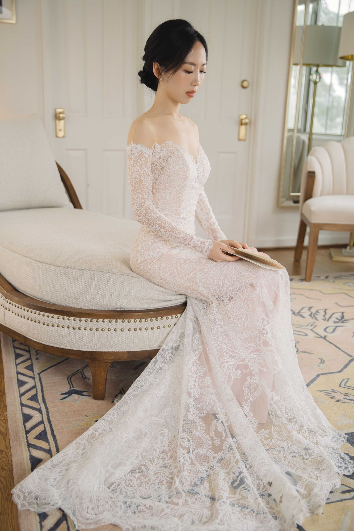Elegant bride in white lace dress sitting gracefully on a round chair in a bright room.