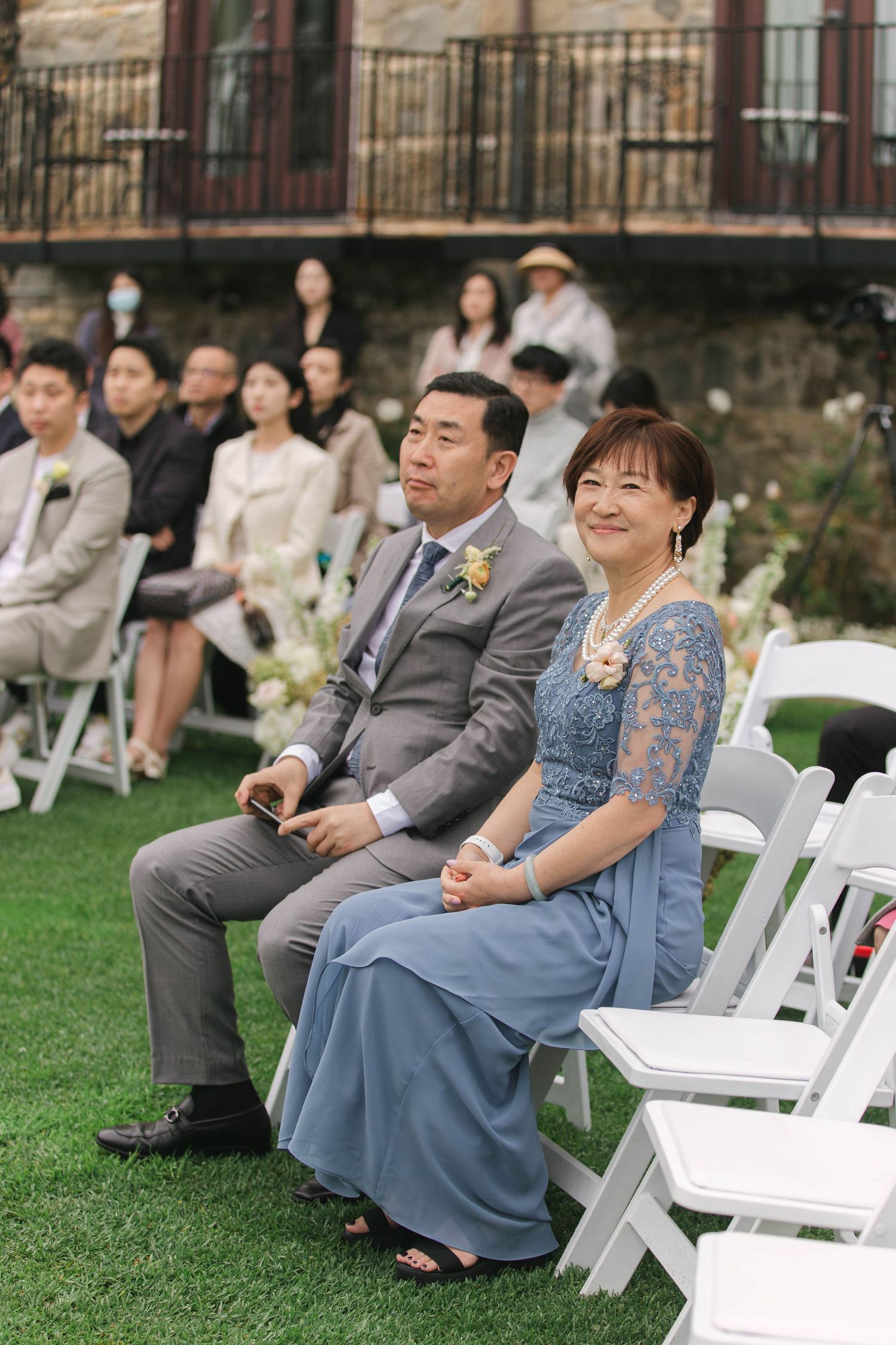 Wedding ceremony guests seated on white chairs on a lawn in formal attire.