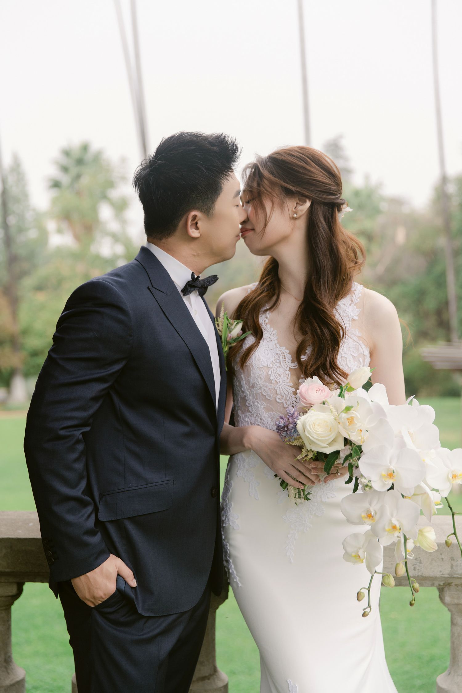 A romantic wedding couple shares a kiss in a garden setting with white floral bouquet.