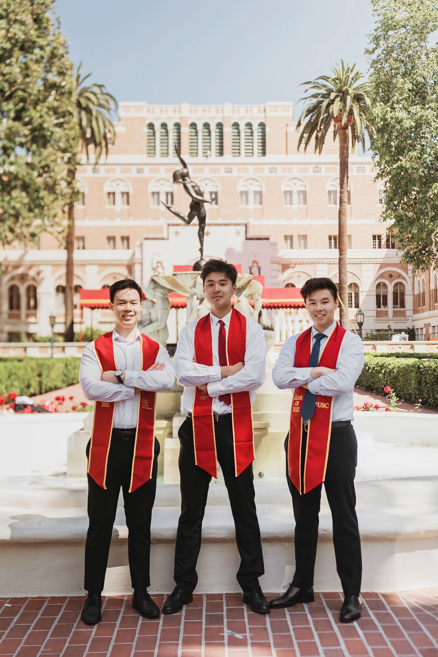 Group of graduates in red stoles strike playful poses in front of historic university building.