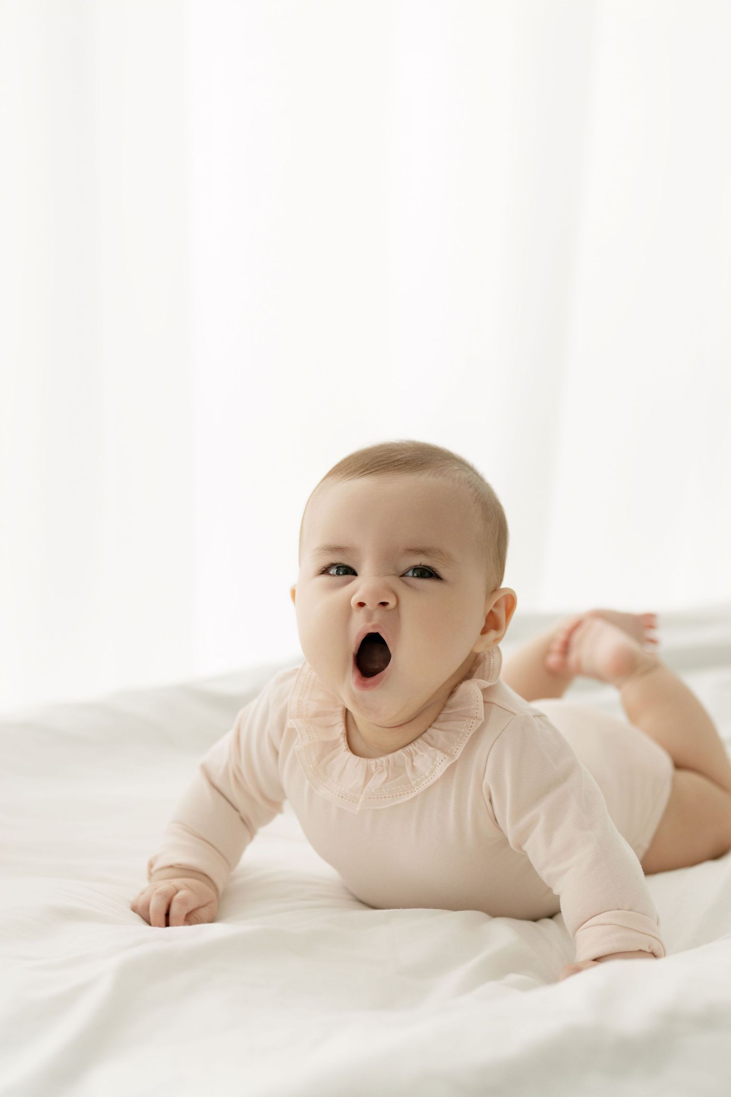 Baby in cream onesie lying on white bedding making expressive faces.