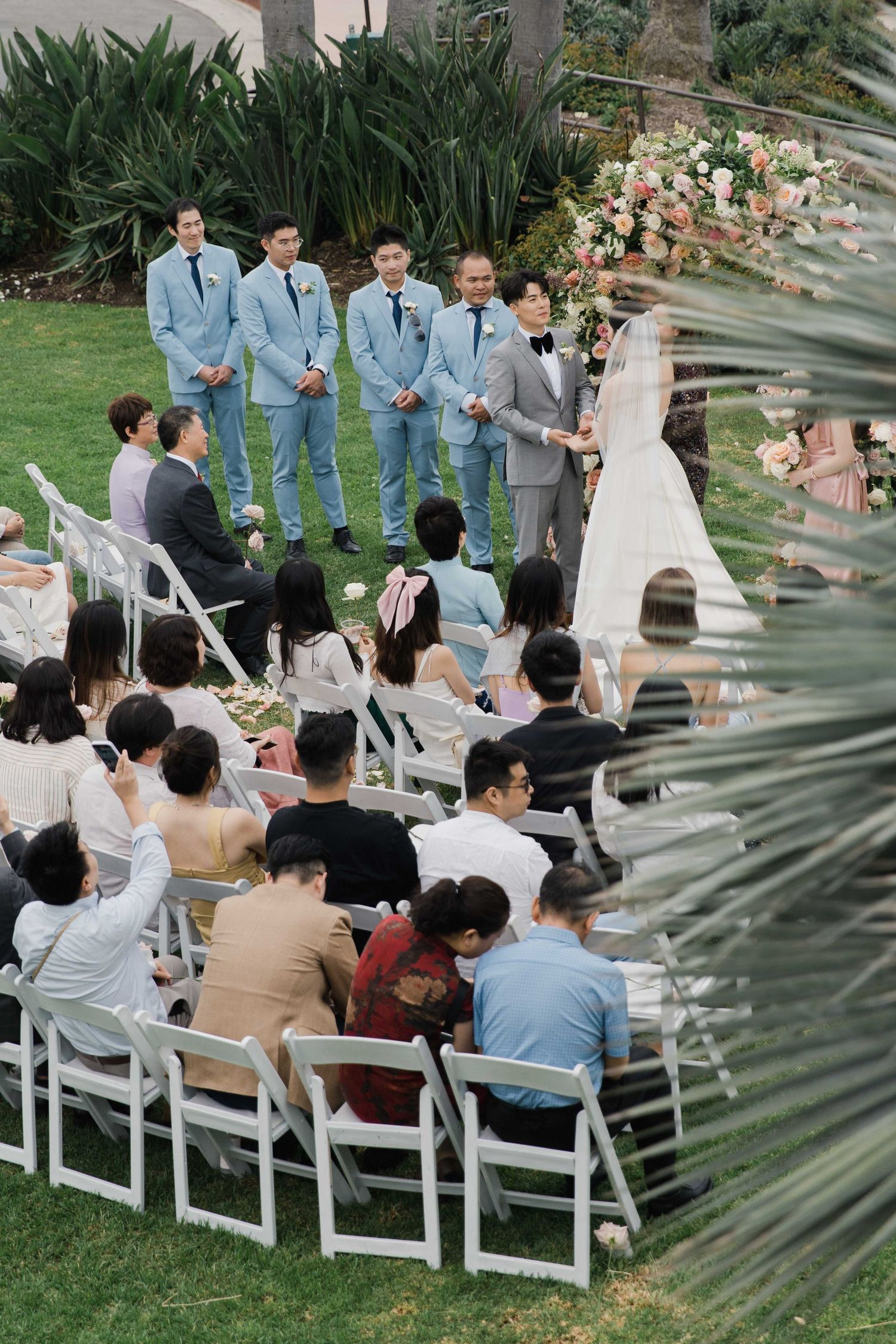 Outdoor wedding ceremony with groomsmen in light blue suits and guests seated on white chairs.