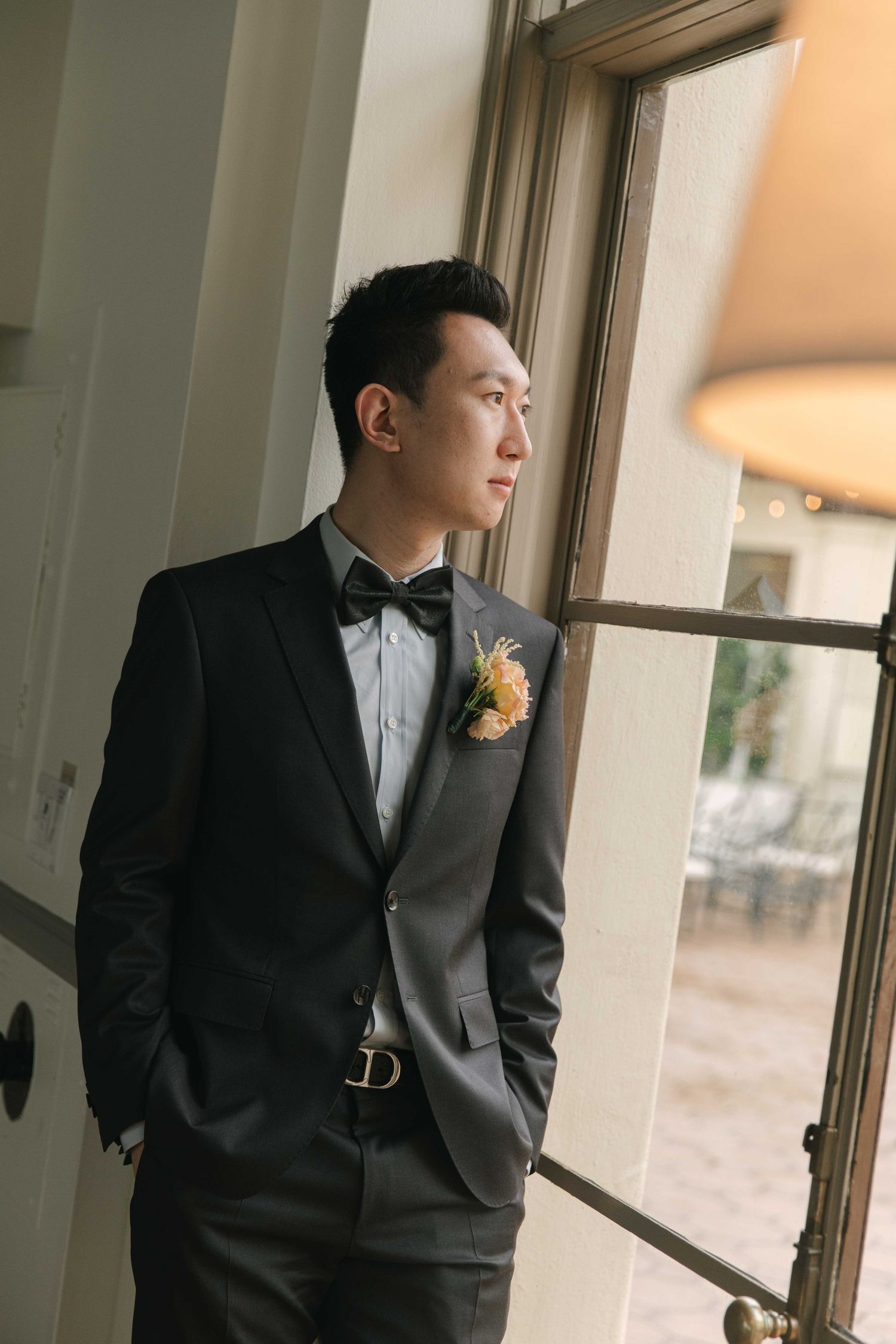 Person in a black tuxedo poses thoughtfully by a window in natural light.
