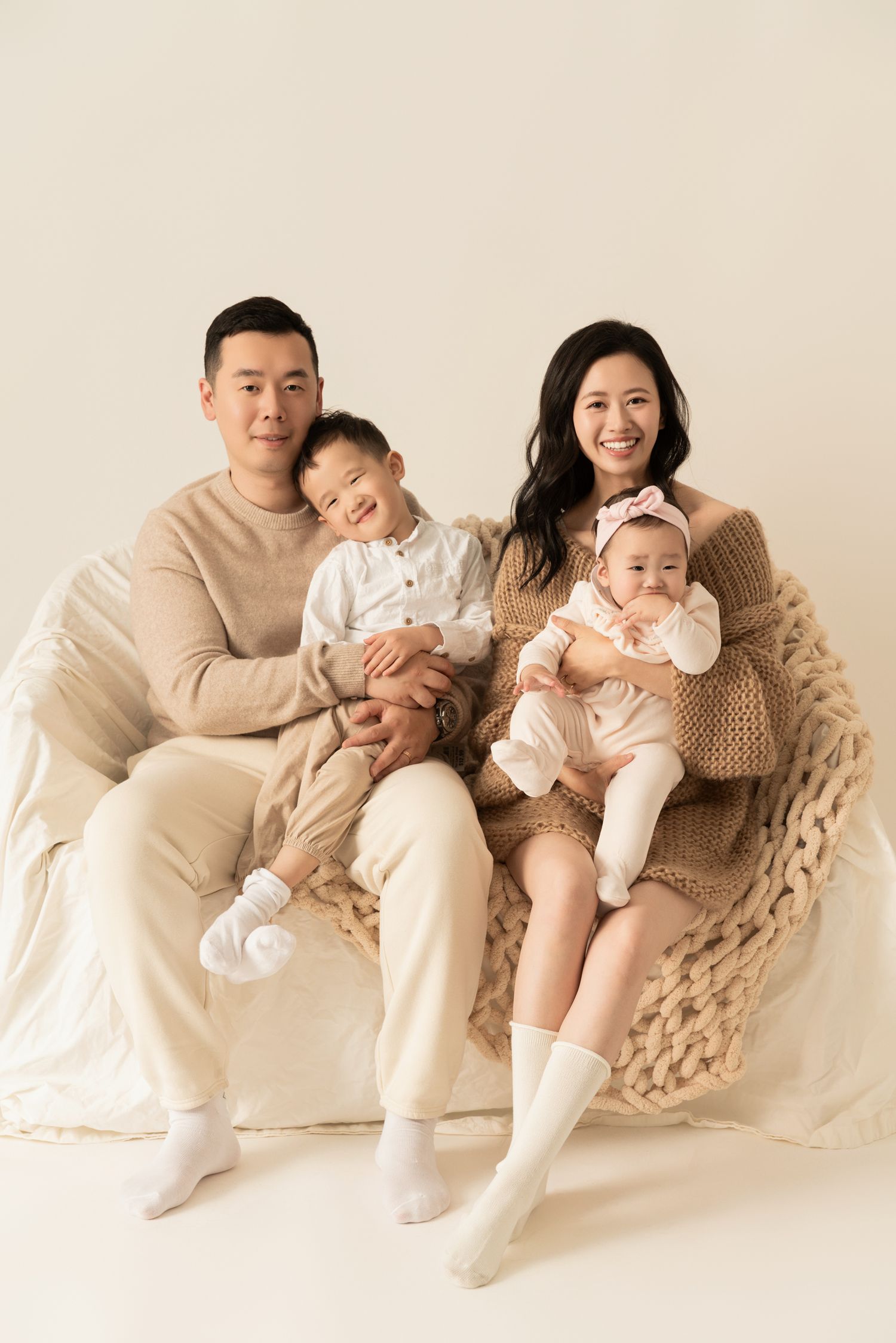 Family portrait of four people sitting together on a beige couch in matching neutral tones.