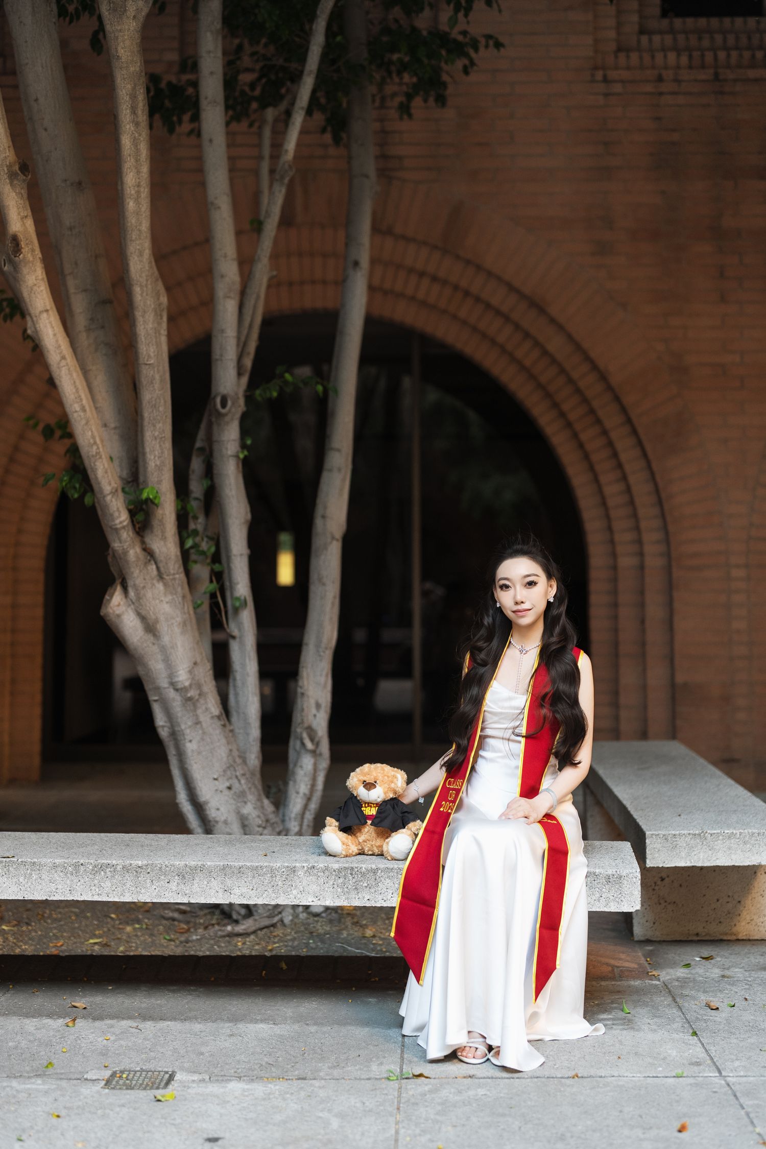 Graduate in white dress poses near brick building with arched walkways.