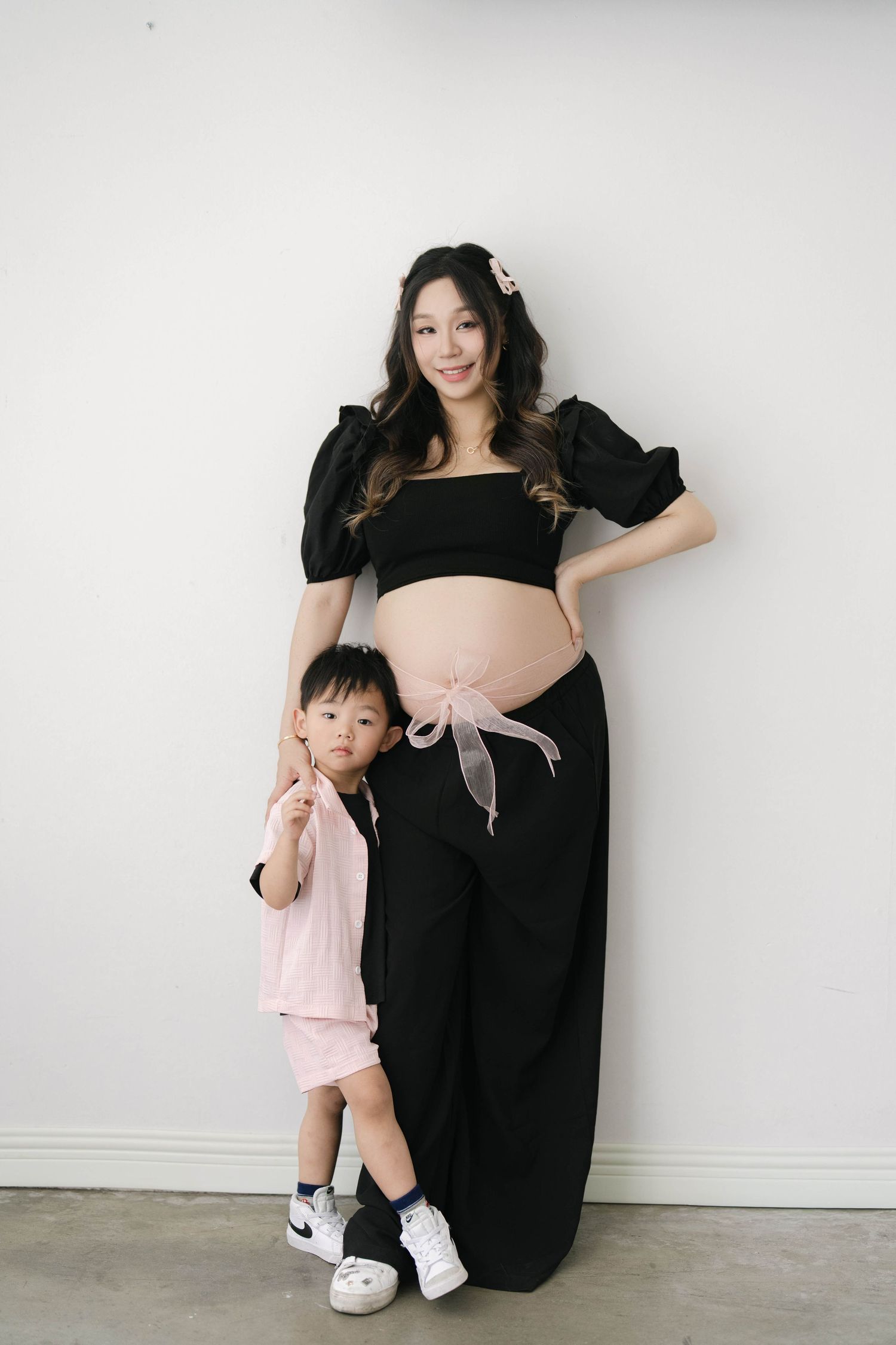 Mother in black dress poses with child in pink outfit against white wall for maternity photo.