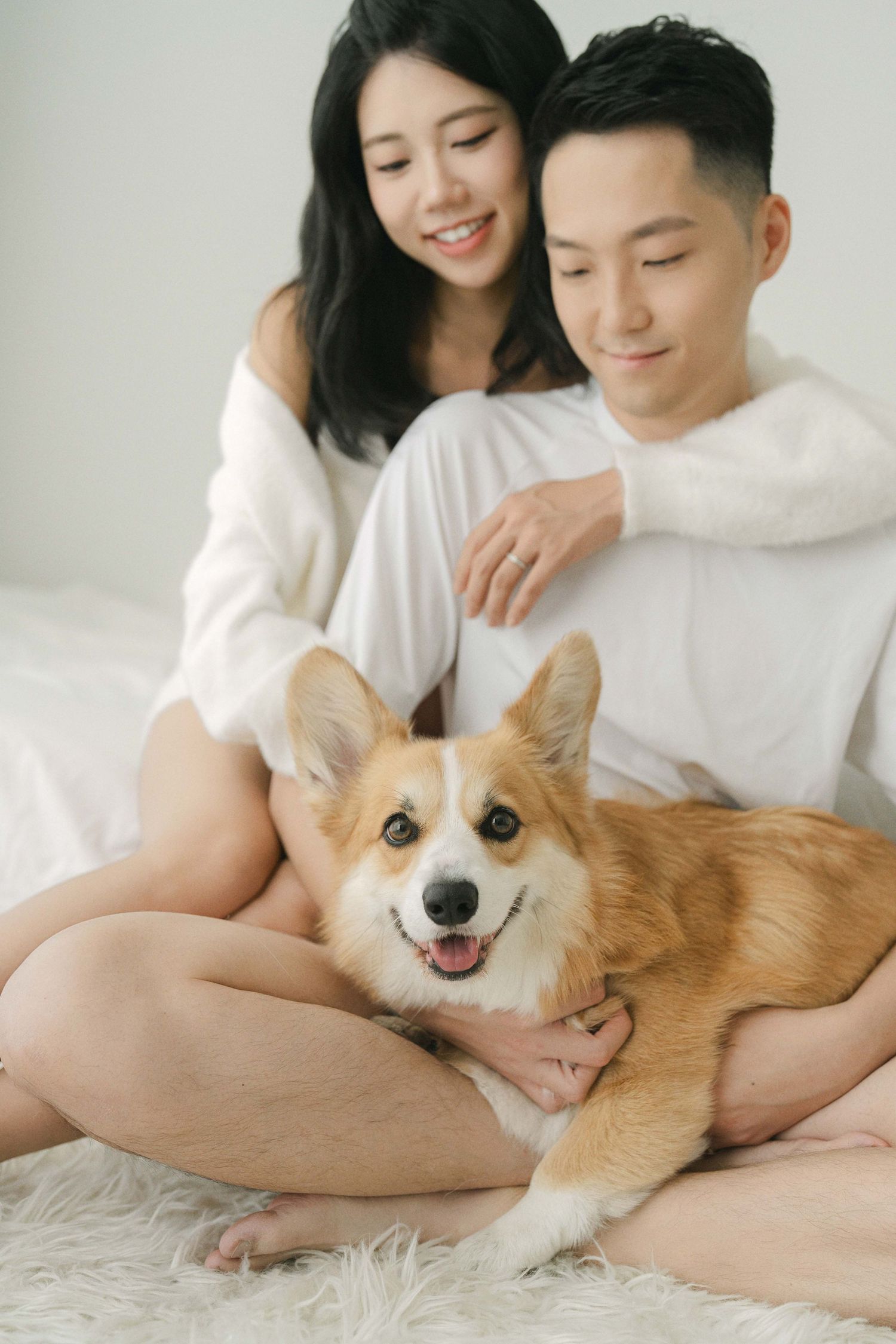 A couple relaxes on white bedding with their smiling Corgi dog.